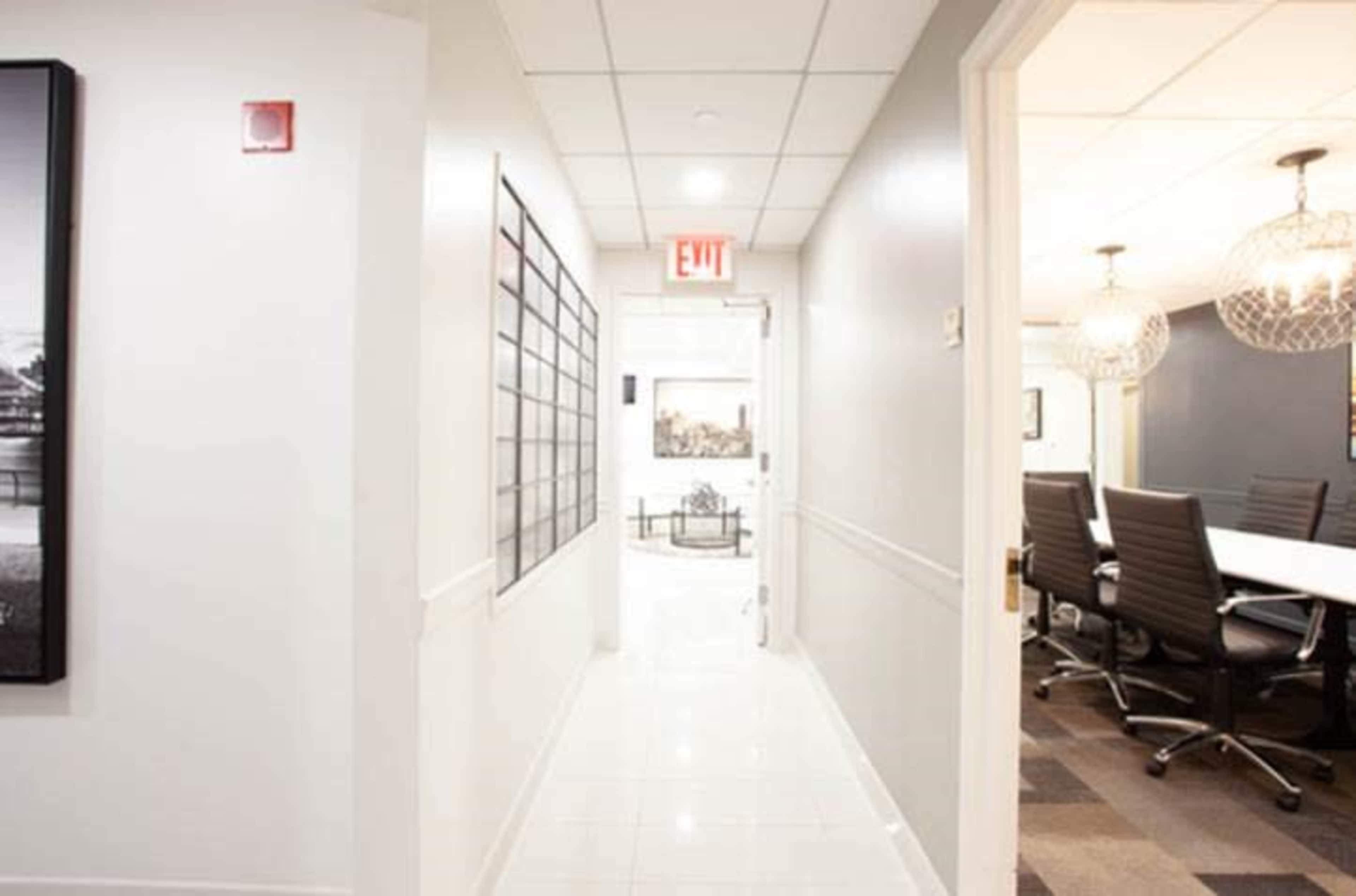A well-lit corridor features white walls, grey flooring, and an exit sign, leading to a meeting room at the end.