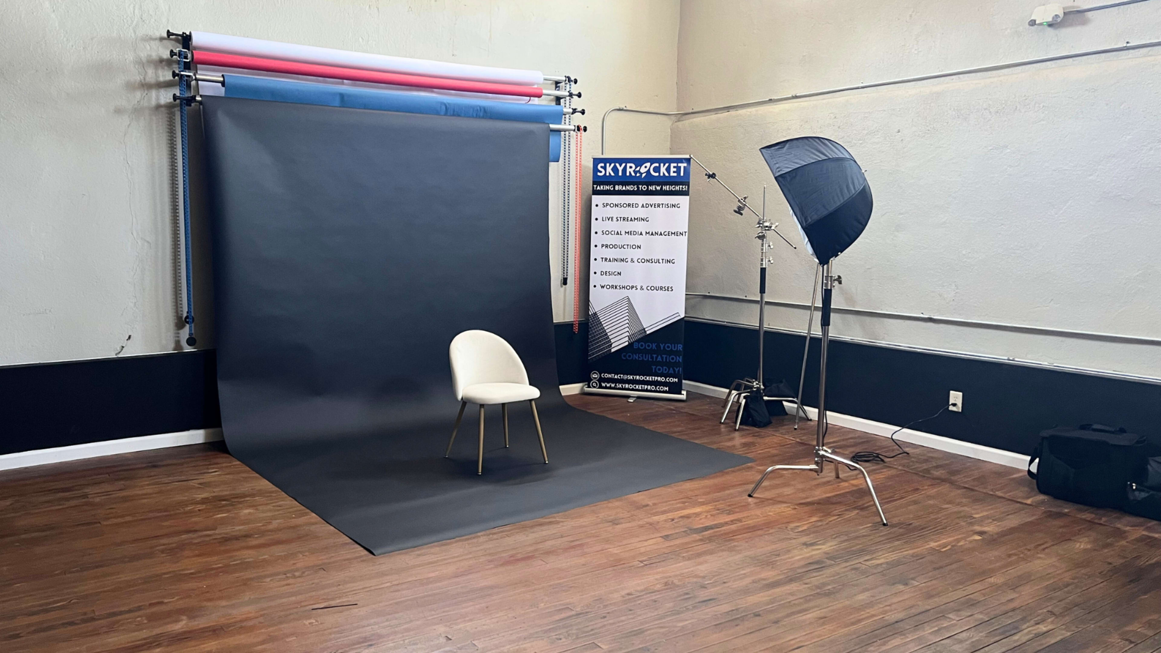 A photography studio setup with a black backdrop, a single chair, and a light source on a stand, near a promotional banner.