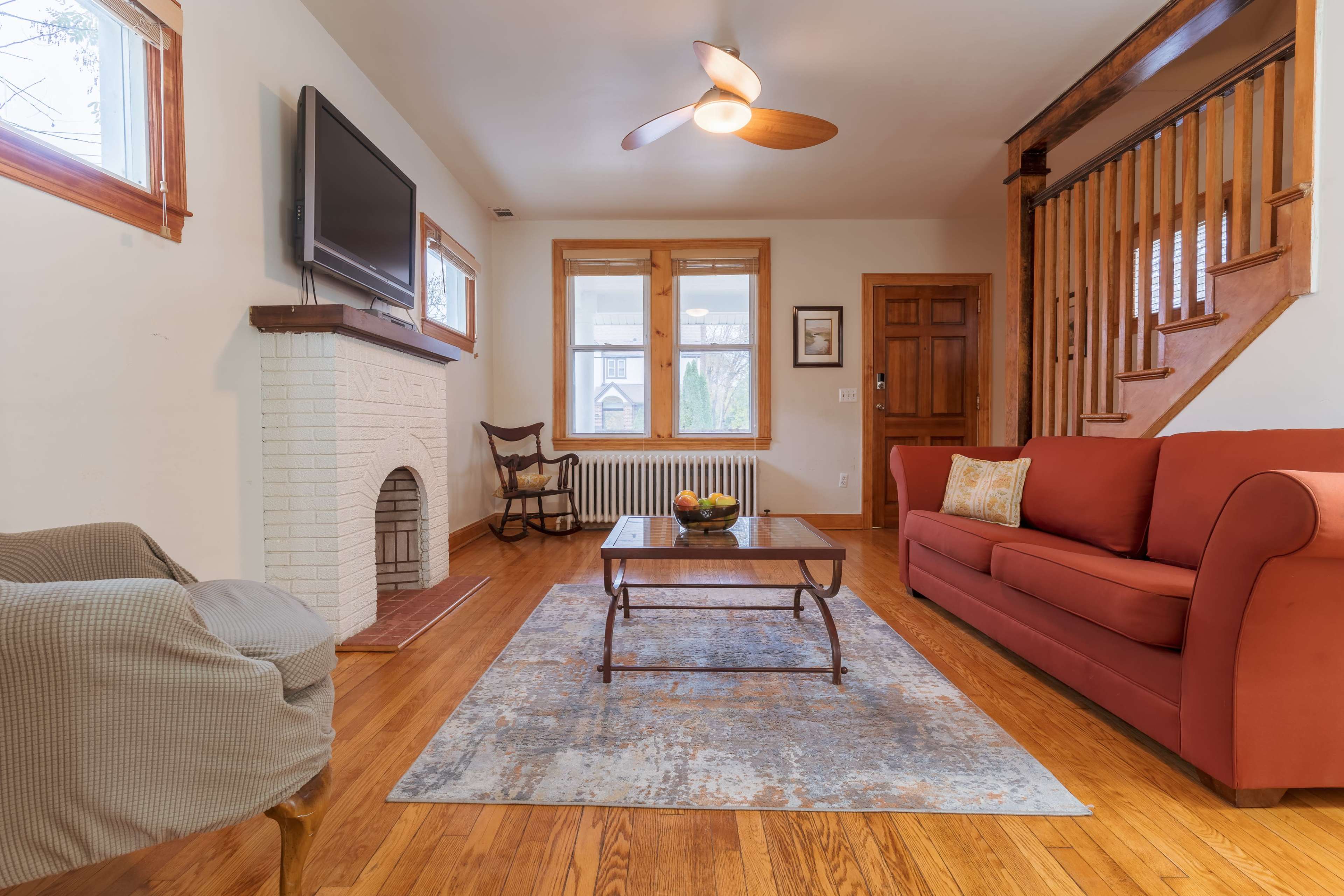 The living room features a red sofa, a wooden coffee table, and a modern ceiling fan, with an inviting fireplace and staircase in the background.