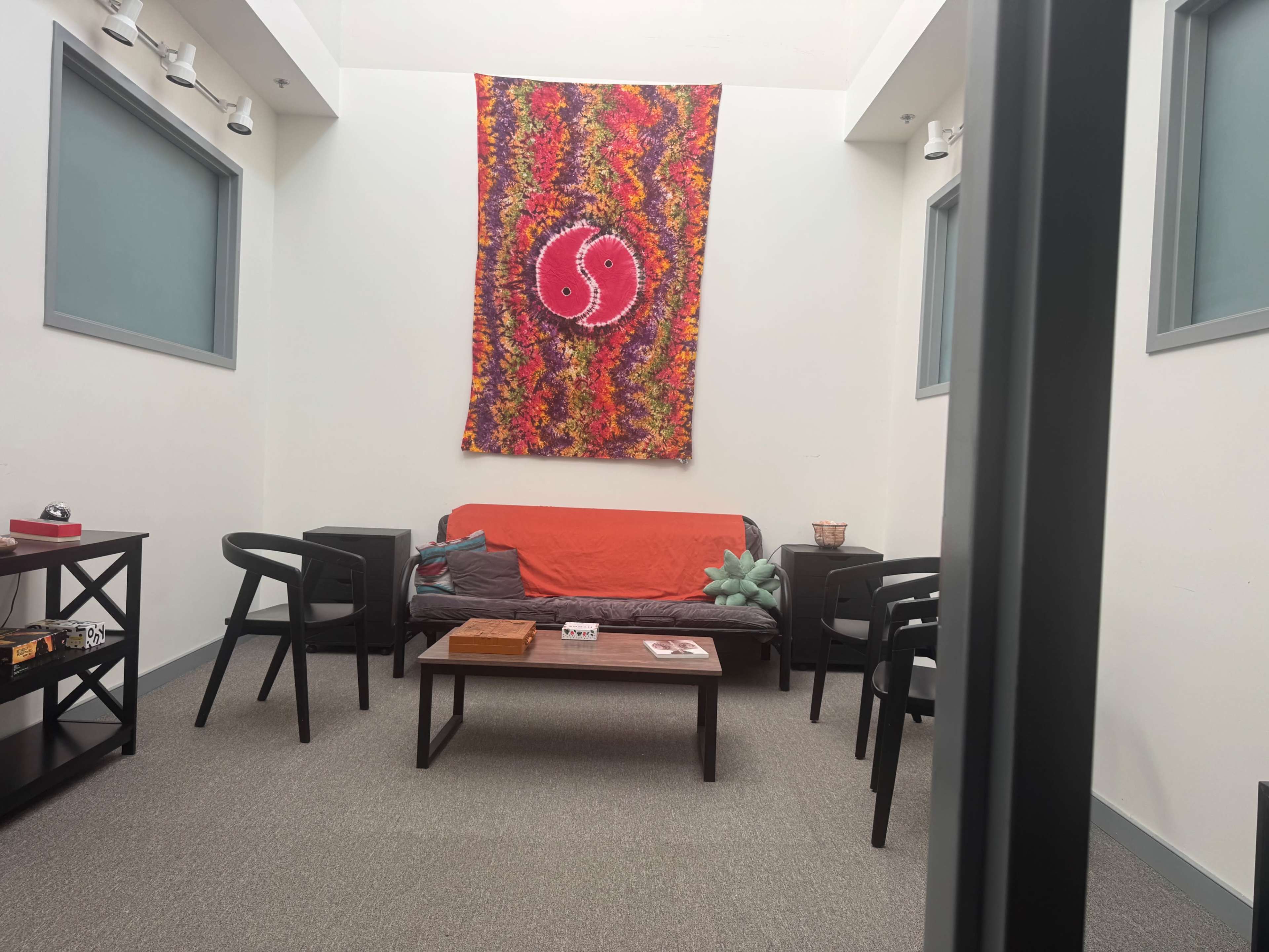 The image shows a small, brightly colored room with seating arrangements including a black sofa and chairs, a coffee table, and a decorative wall hanging.