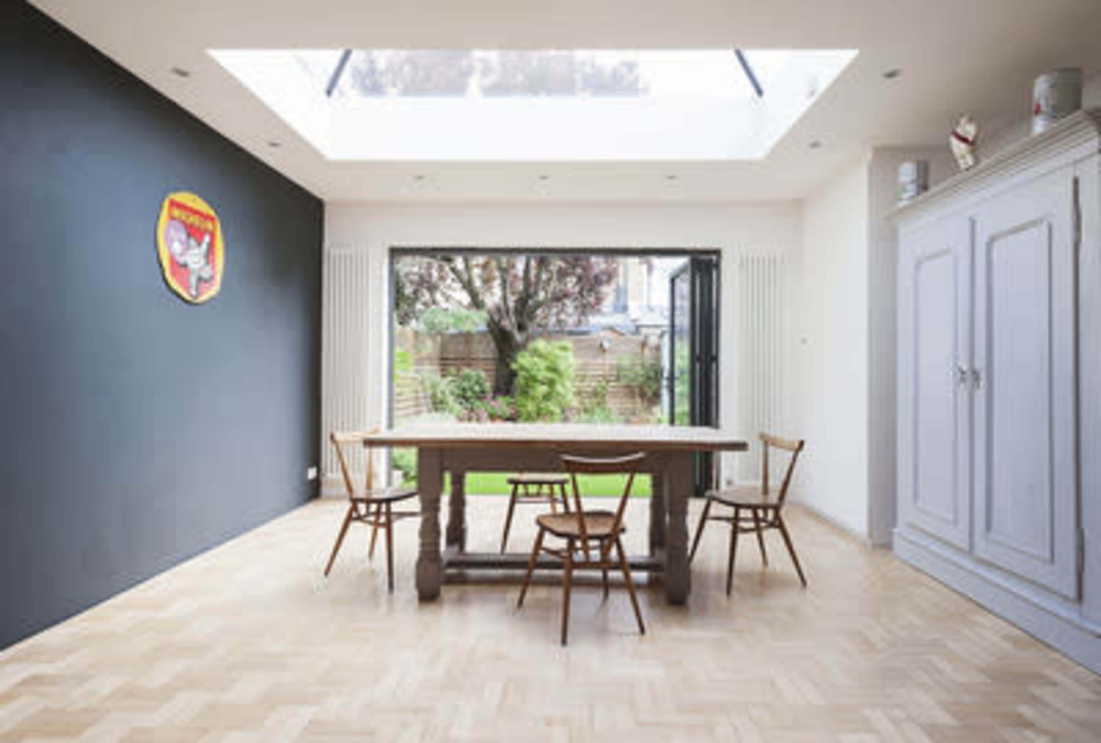 A minimalist dining area features a large wooden table surrounded by four chairs, with a skylight above and a view of a garden through open glass doors.