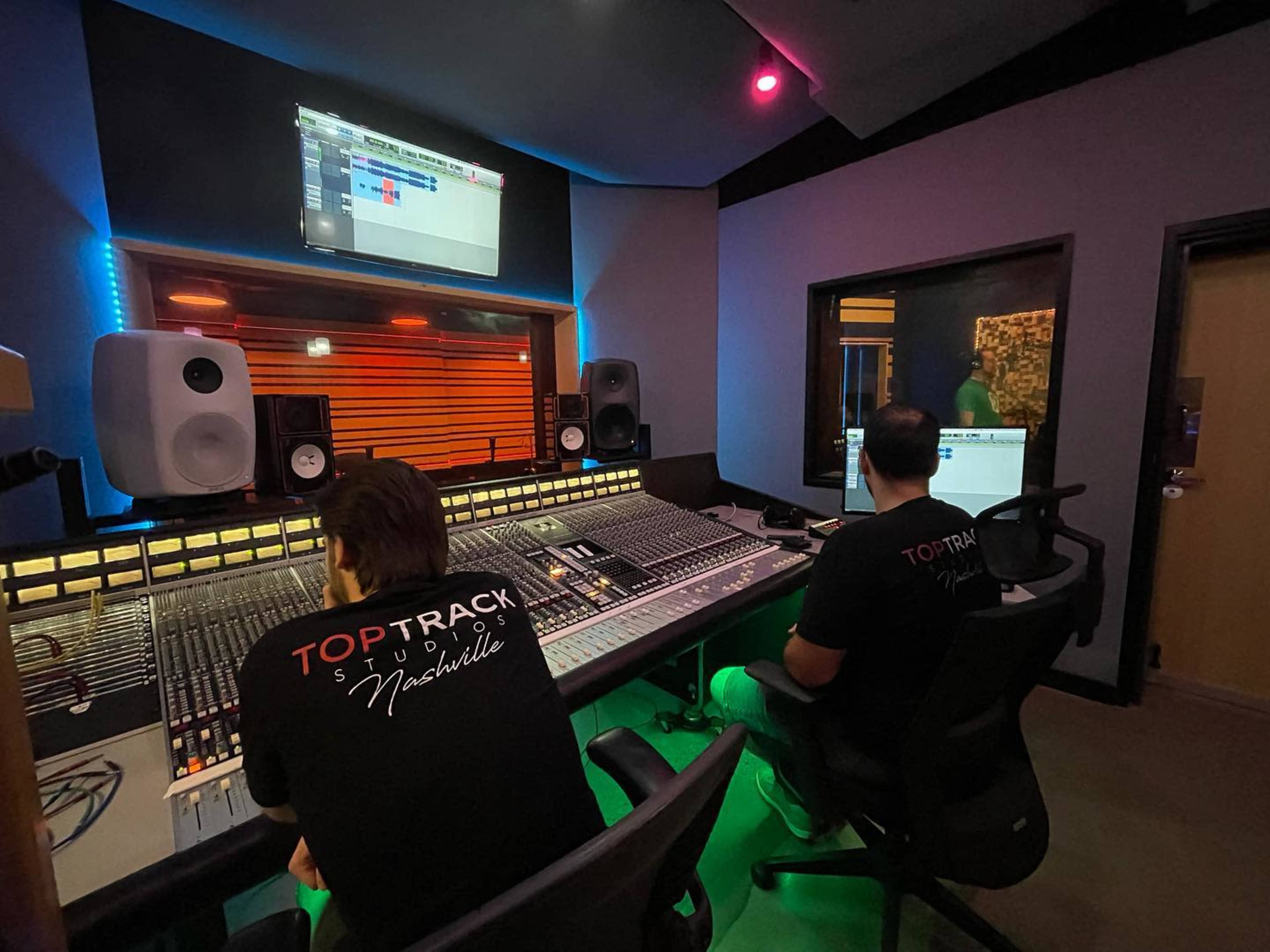 Two sound engineers work at a recording studio with a large mixing console and monitors, focused on a digital audio workstation screen.