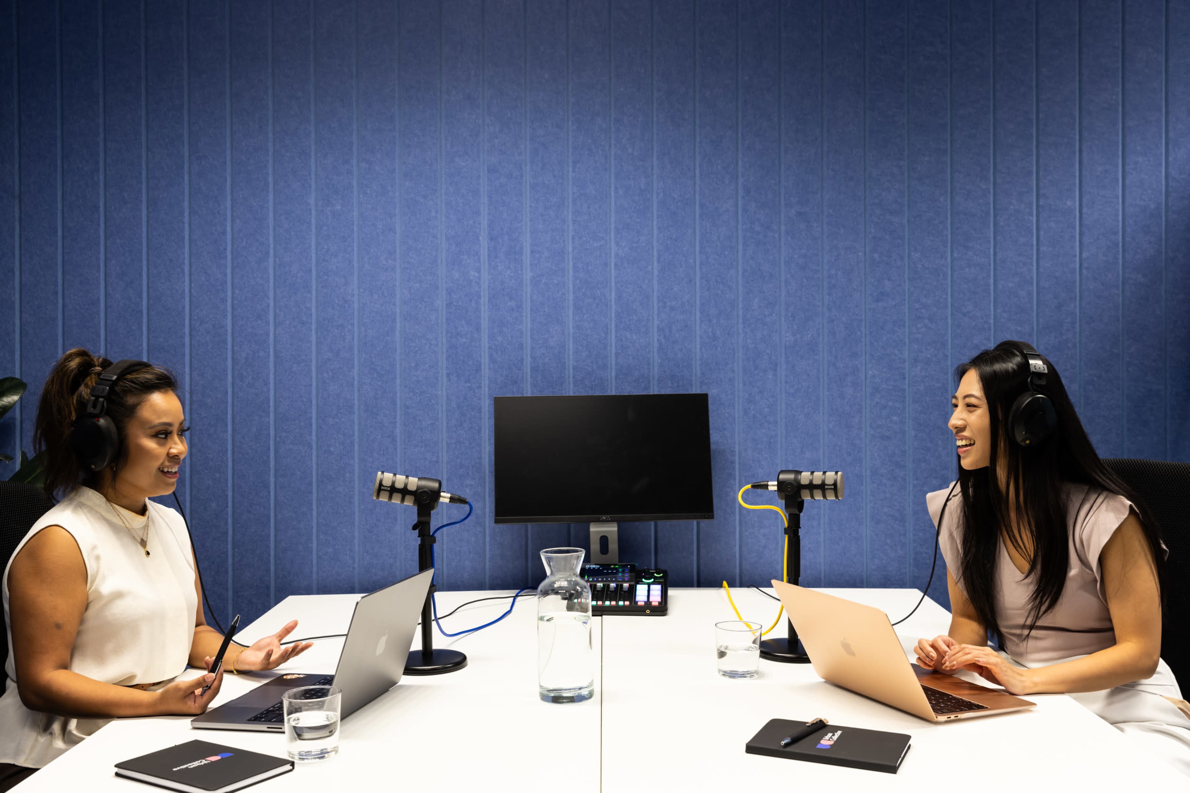 Two women are seated at a table with laptops and microphones, engaged in conversation in a blue-walled podcast recording studio.