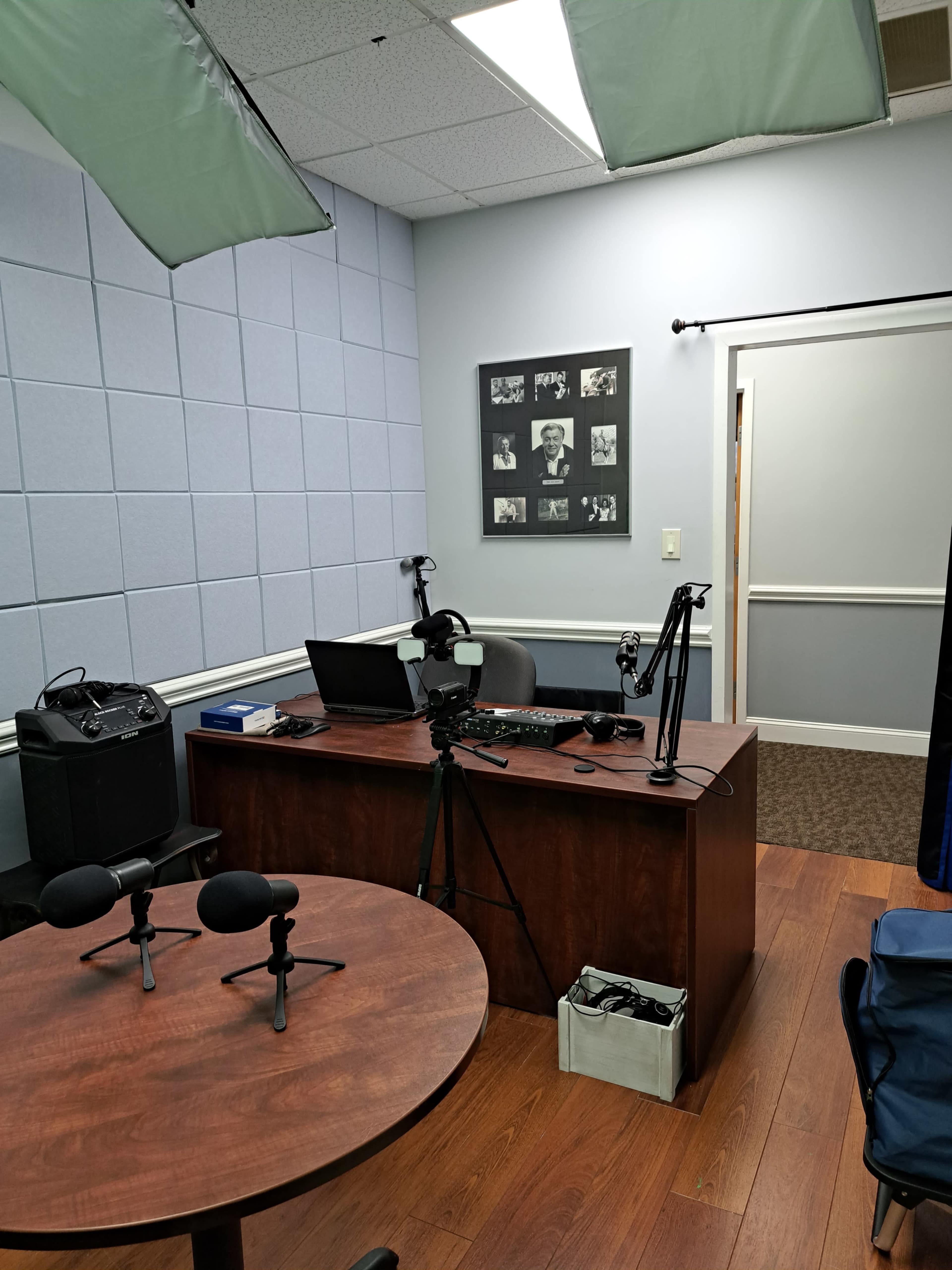 A small video recording studio featuring a wooden desk with a computer, microphones, and a backdrop of black-and-white photos on the wall.