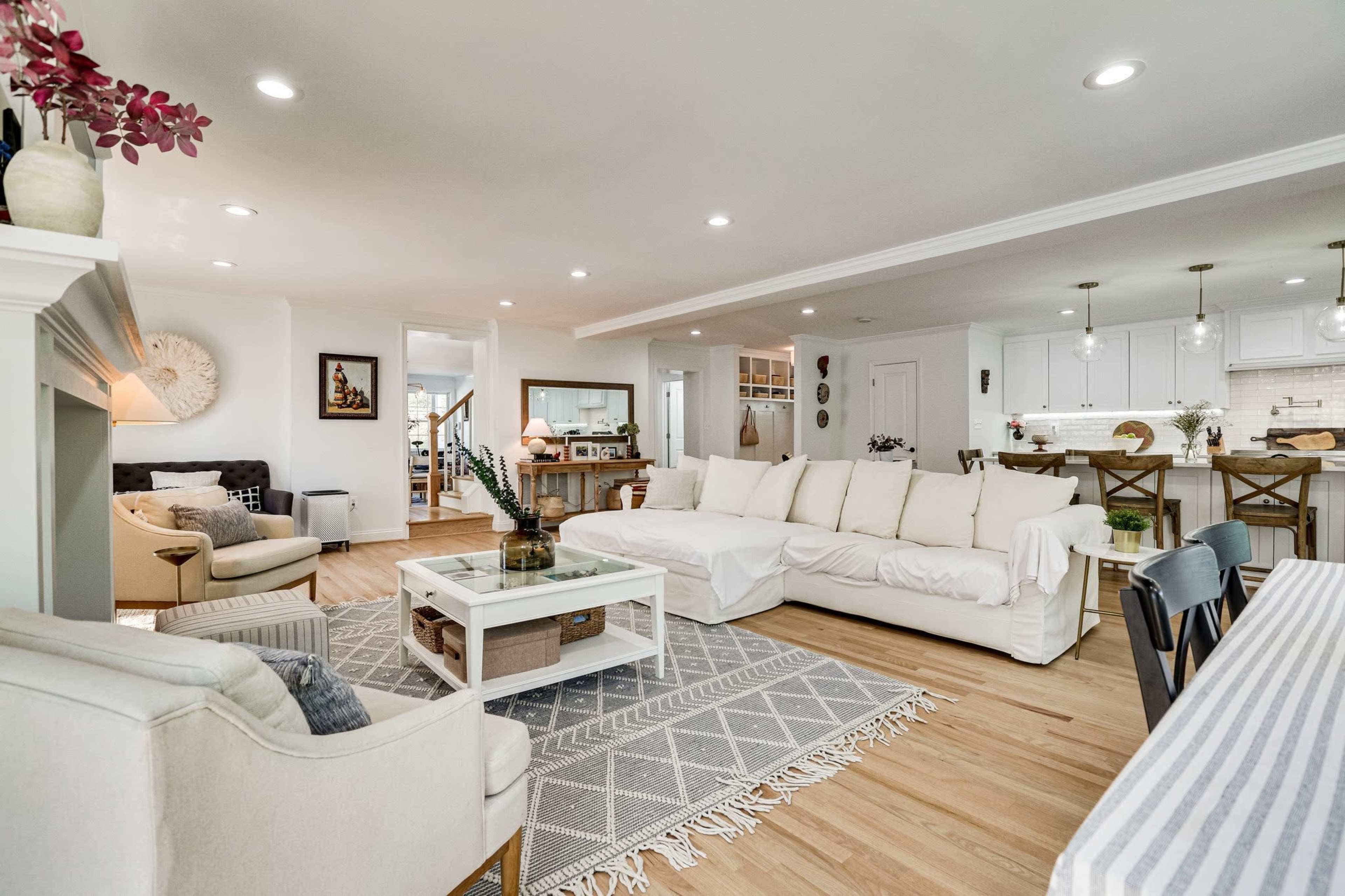 The image shows a spacious, modern living area featuring a large white sectional sofa, a coffee table, and a dining area in the background with white cabinetry and pendant lights.