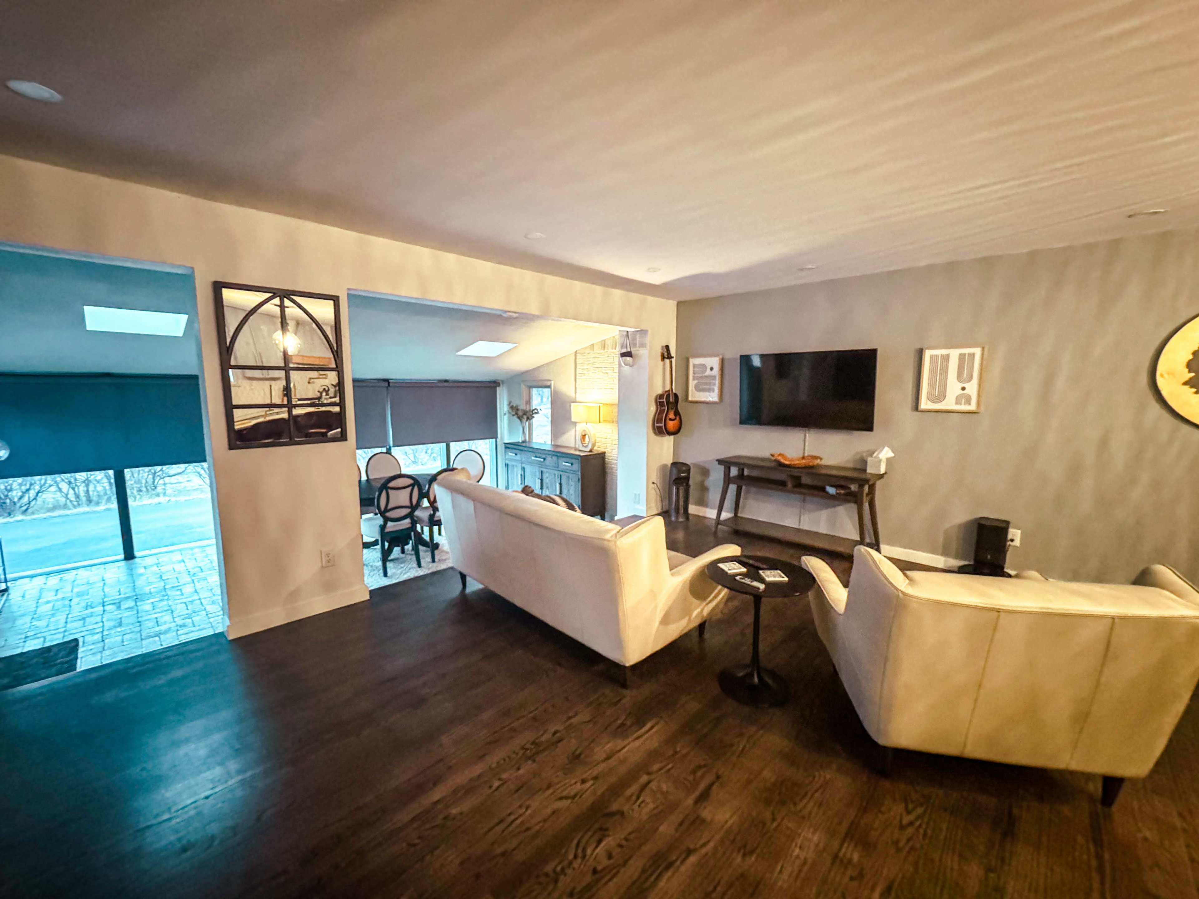 The image shows a modern living room featuring two white couches, a small black table, and a TV mounted on the wall, with a dining area visible in the background.