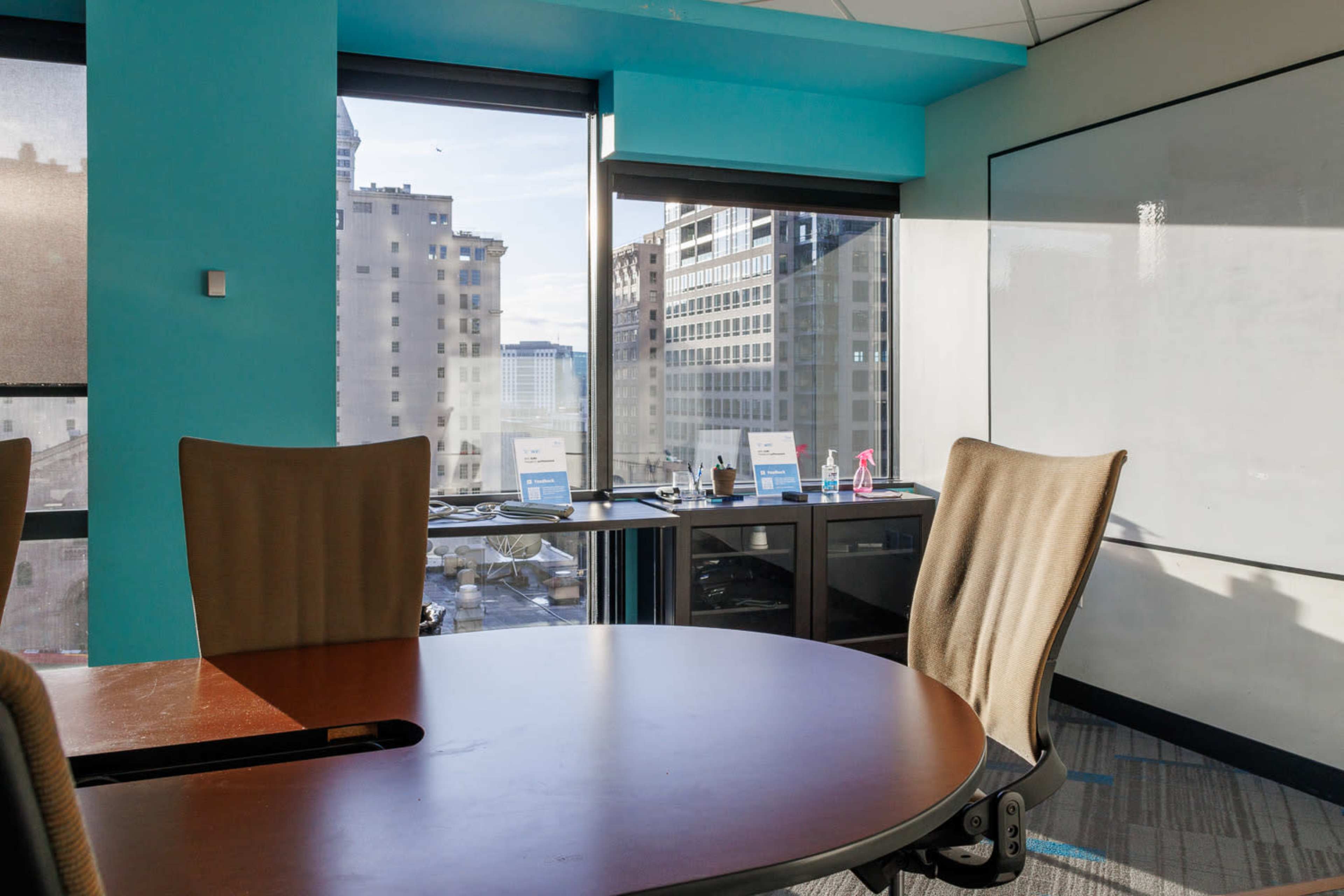 A conference room features a round wooden table, three beige chairs, and large windows showcasing a city skyline.