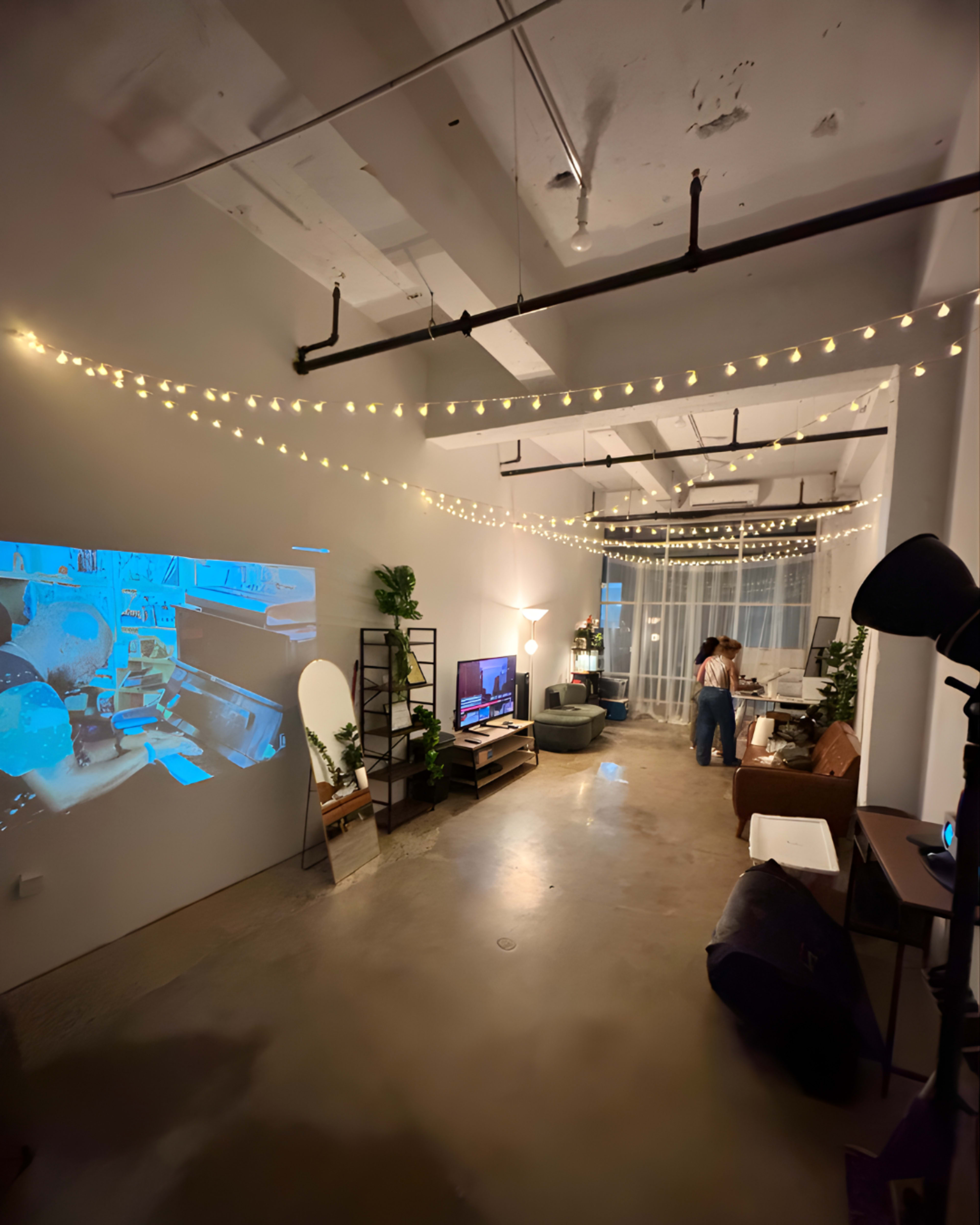 A modern living room features string lights overhead, a television against one wall, and a person standing near a desk with plants and a wall projection displaying images.
