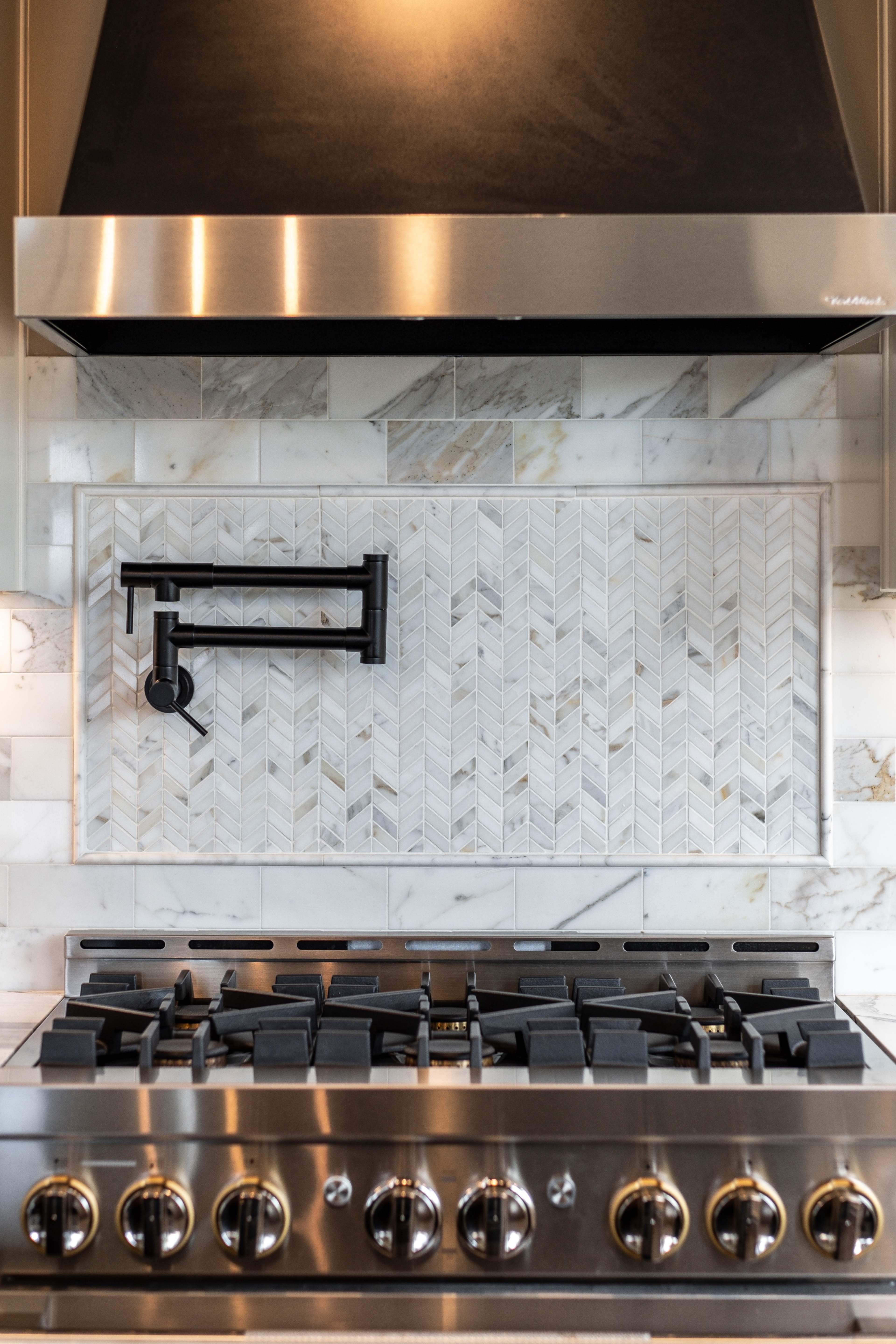 The image shows a modern kitchen backsplash with a herringbone pattern in marble above a stainless steel gas stove and a metallic range hood.