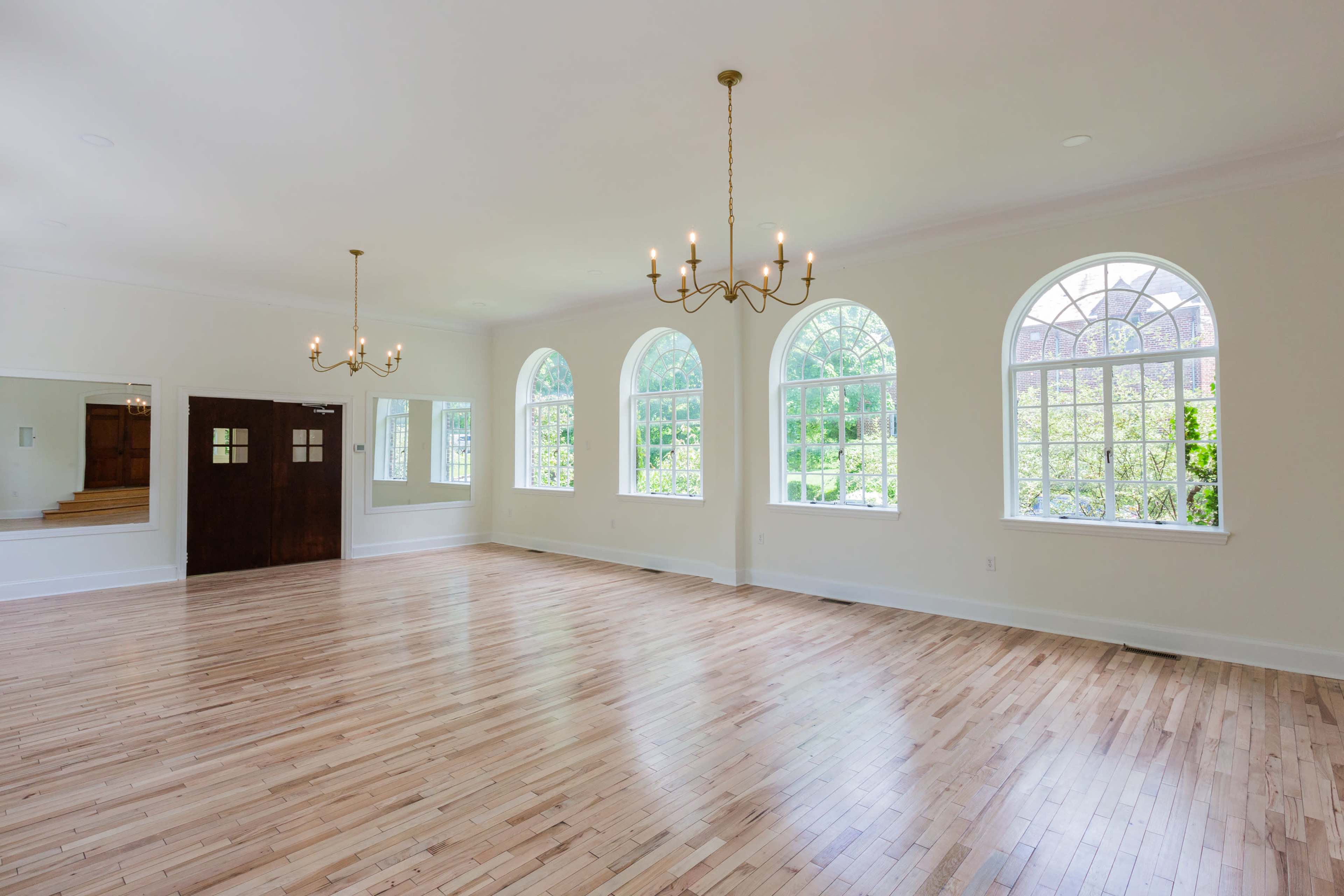An empty room with hardwood flooring and large arched windows allowing natural light to enter, along with a chandelier hanging from the ceiling.
