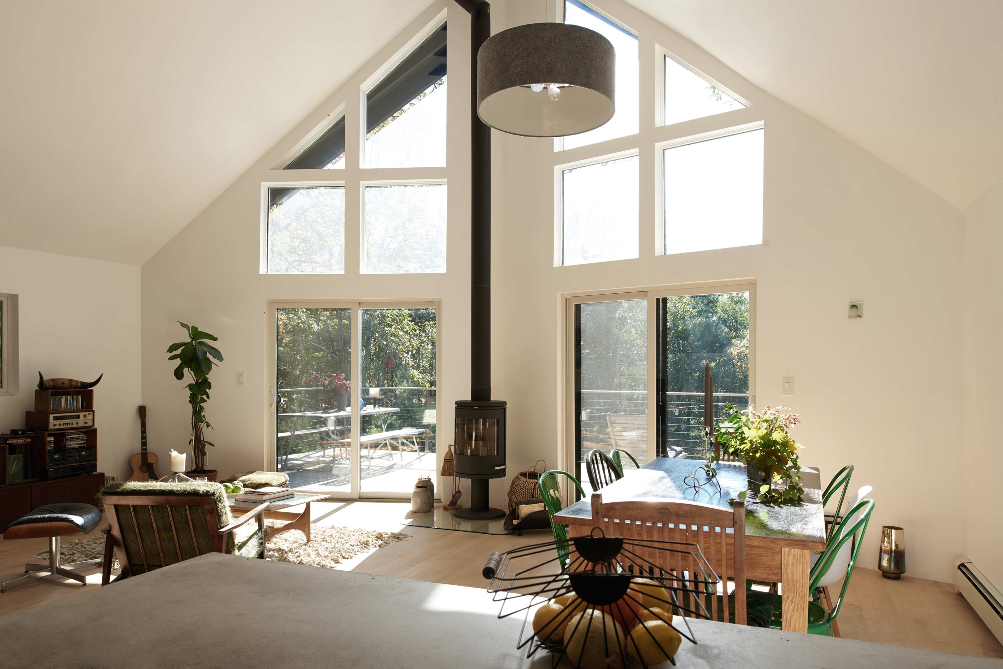 A modern living space with tall windows, a wood stove, and a dining area furnished with a table and colorful chairs.