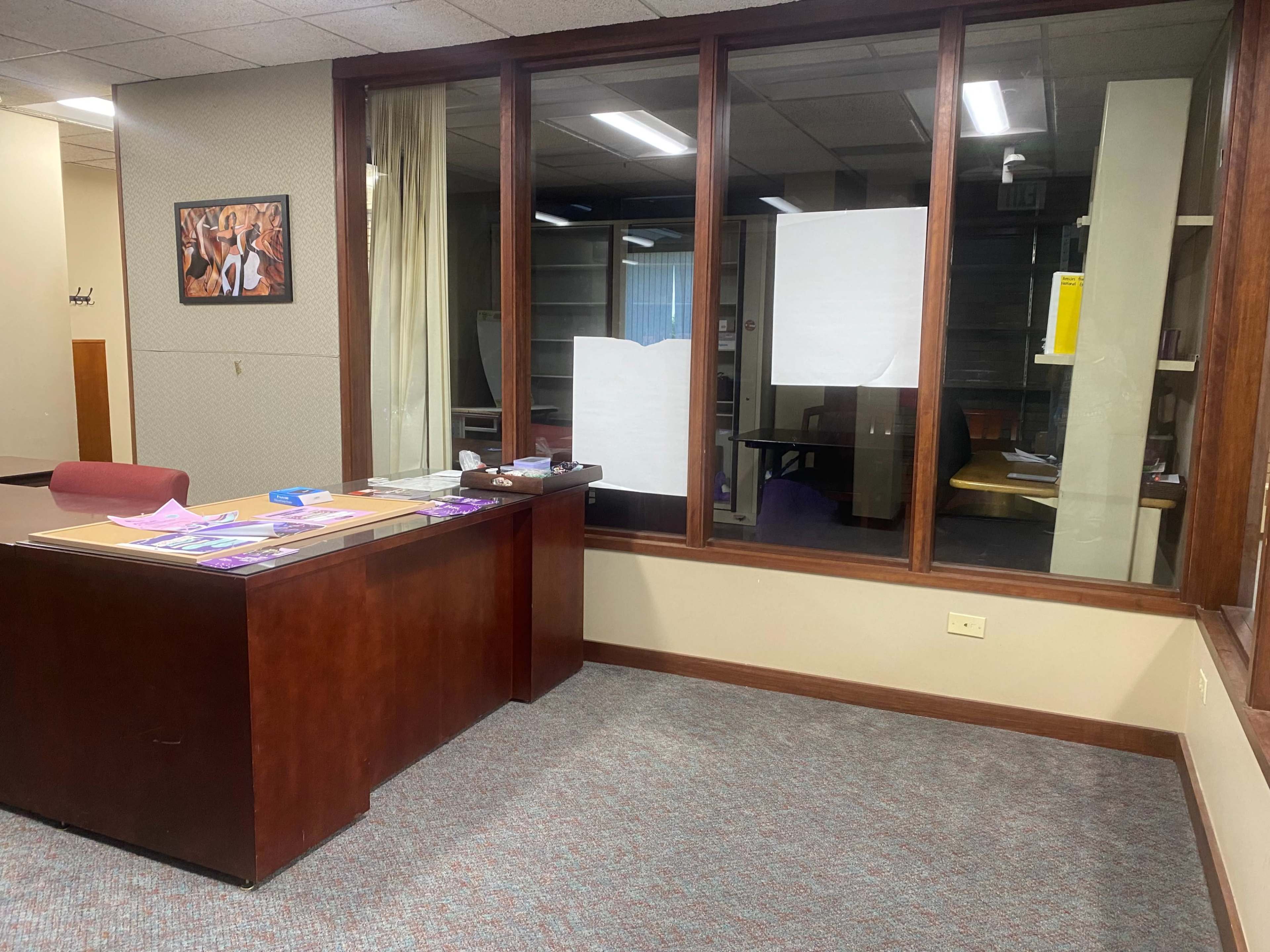 An office space features a brown desk with paperwork and notices, next to large windows looking into an empty meeting area.