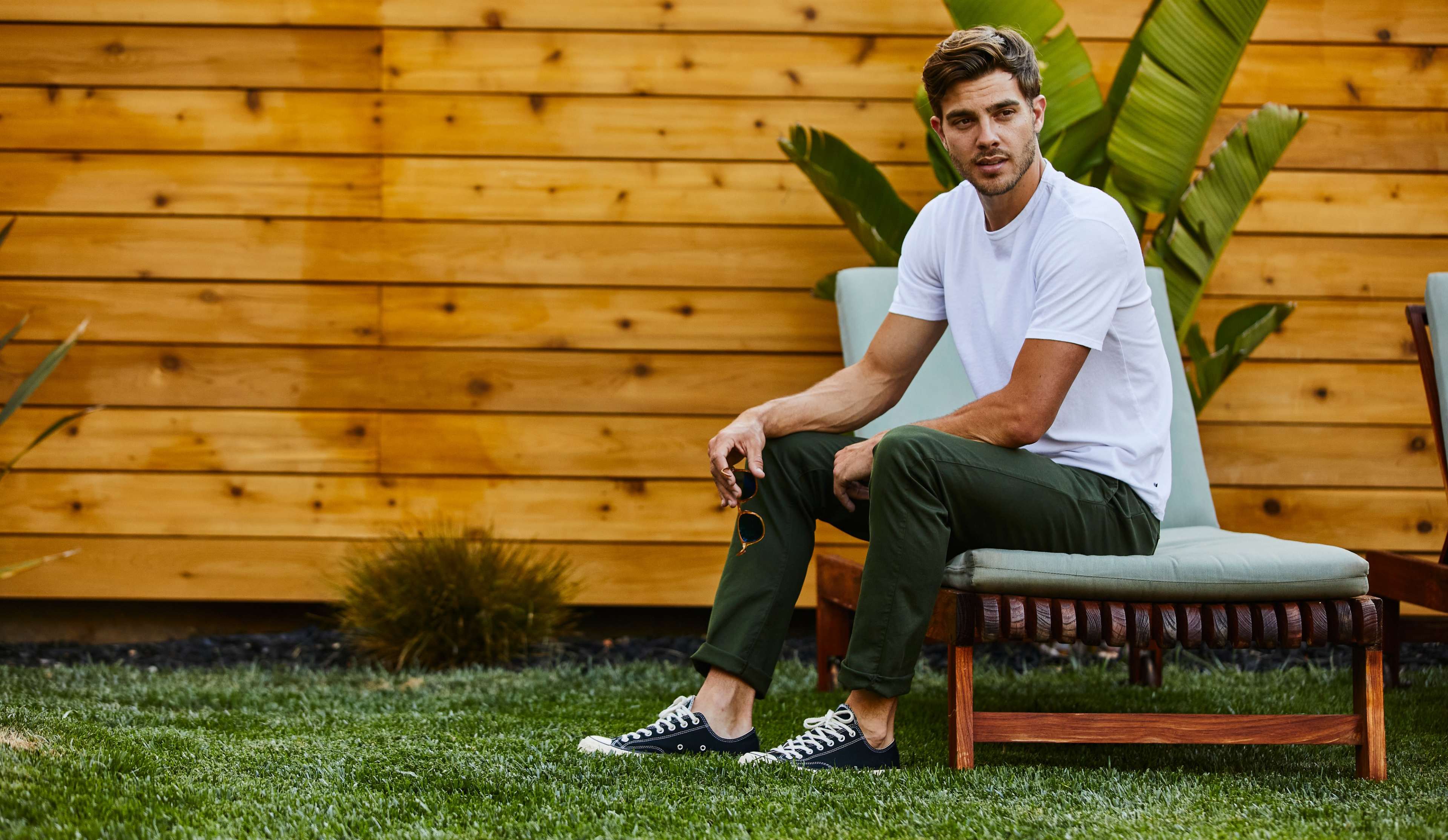 A man sits on a lounge chair in a grassy area with a wooden wall and tropical plants in the background.