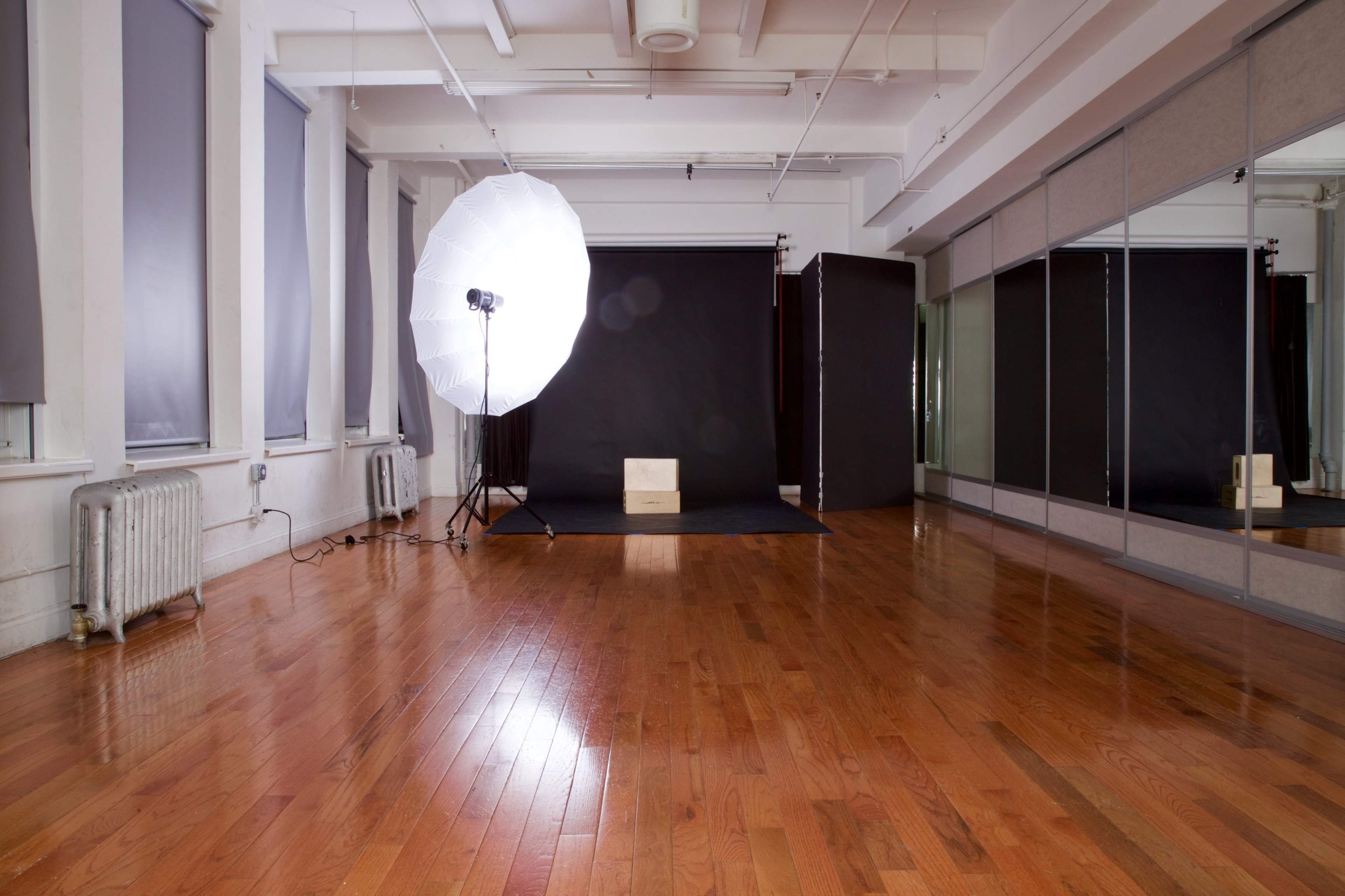 A photography studio features a large softbox light, a black backdrop, and wooden flooring.