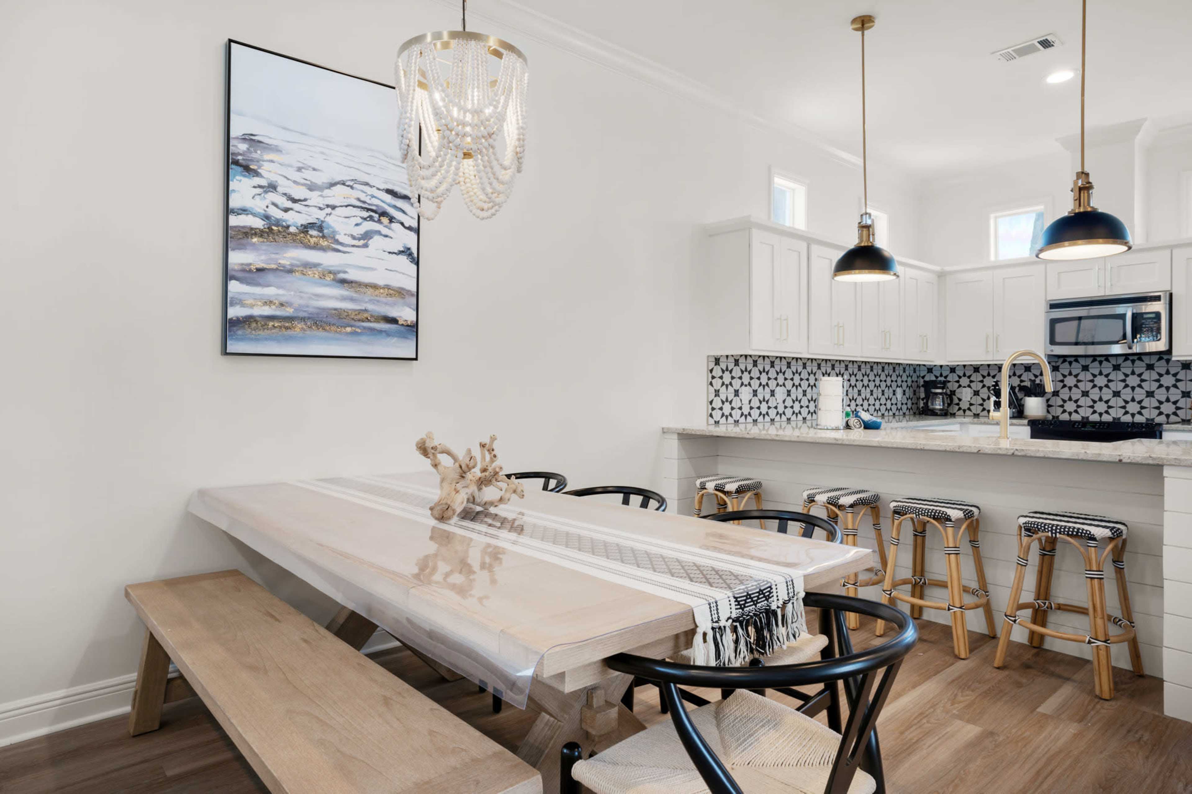 A modern dining area features a long wooden table with a decorative centerpiece, surrounded by sleek black chairs, adjacent to a kitchen with white cabinetry and a patterned tile backsplash.