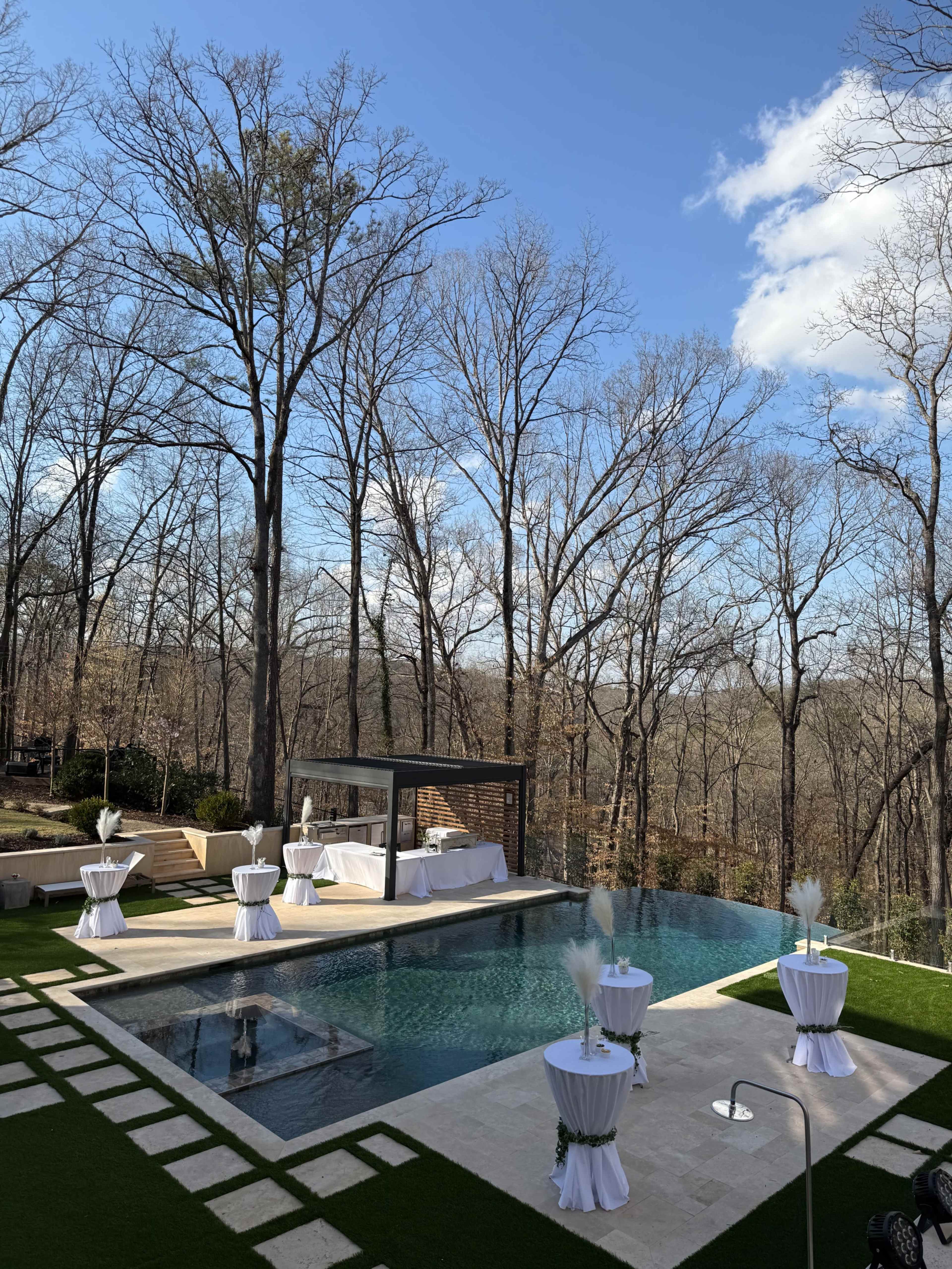 The image shows a spacious outdoor area featuring a rectangular swimming pool, surrounded by several white-topped tables and wooded scenery in the background.