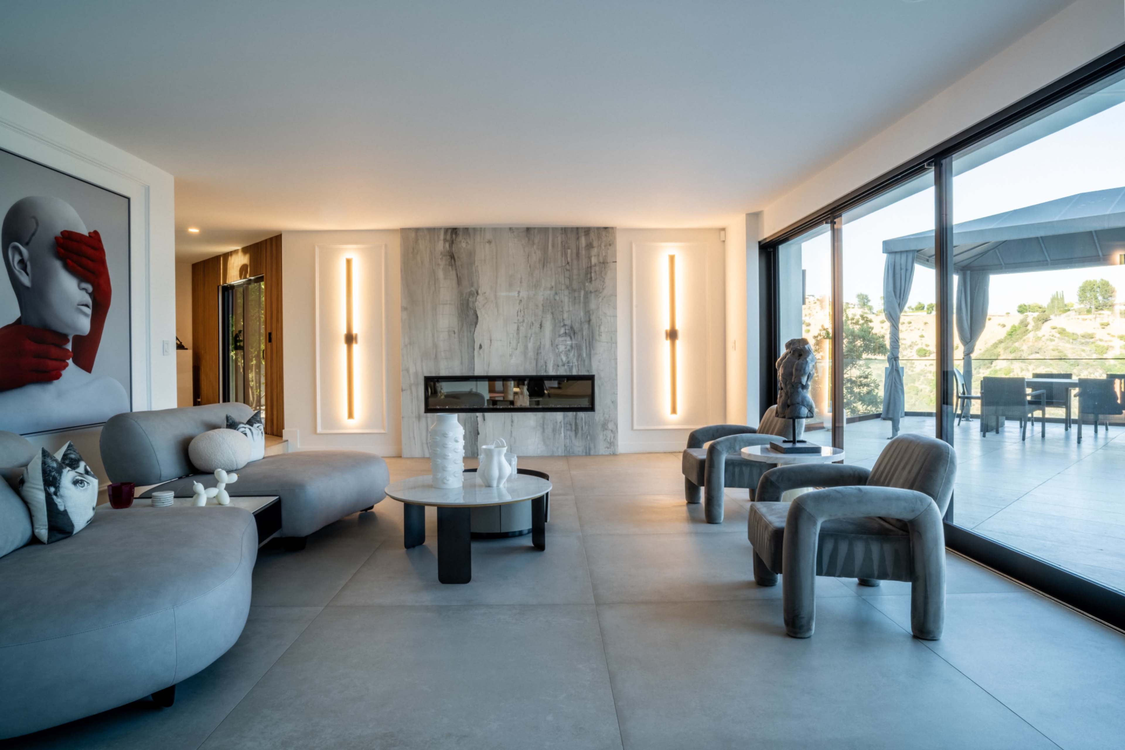 A modern living room features a minimalist design with a large fireplace, sleek furniture, and floor-to-ceiling windows overlooking an outdoor space.