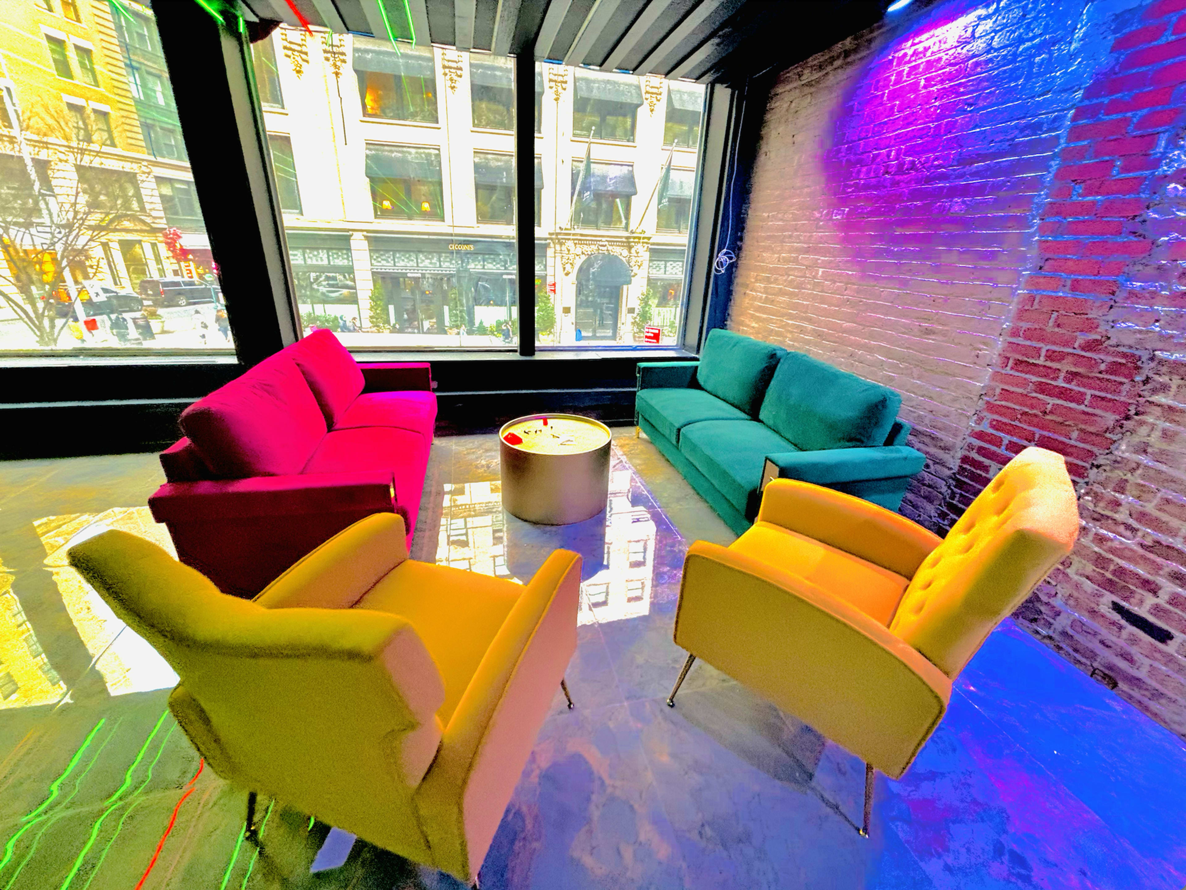A modern lounge area with a circular table surrounded by colorful sofas and chairs, set against large windows displaying a cityscape.