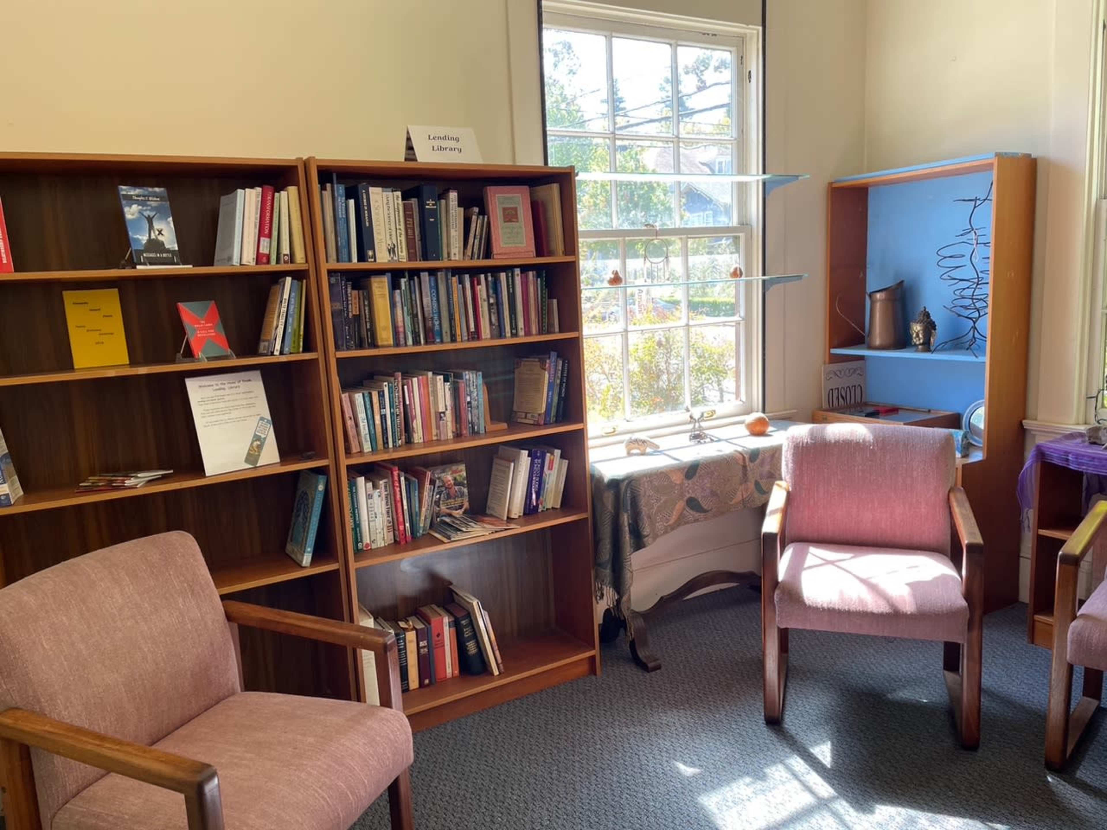 The image shows a small reading room with bookshelves filled with various books, two upholstered chairs facing each other, and a table by a window adorned with decorative items.