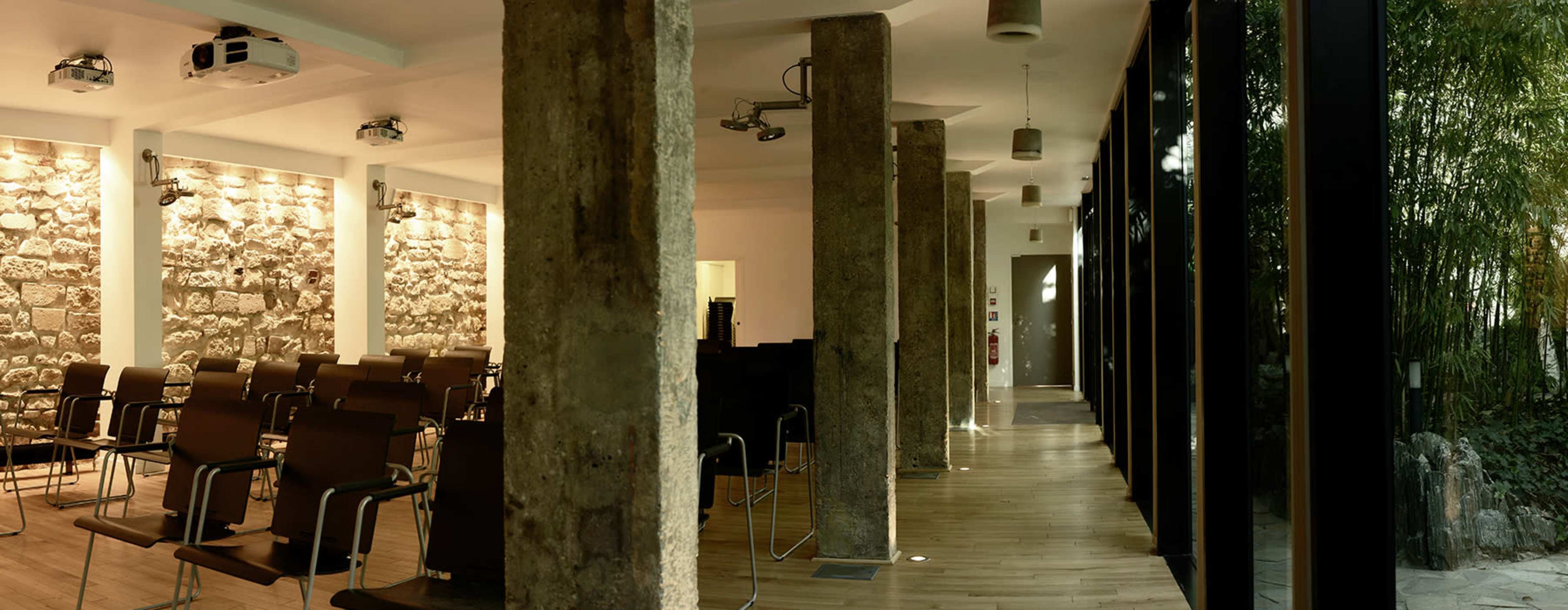 A modern interior space featuring stone walls, wooden floors, and a row of metal chairs facing a projection area, with large windows along one side.