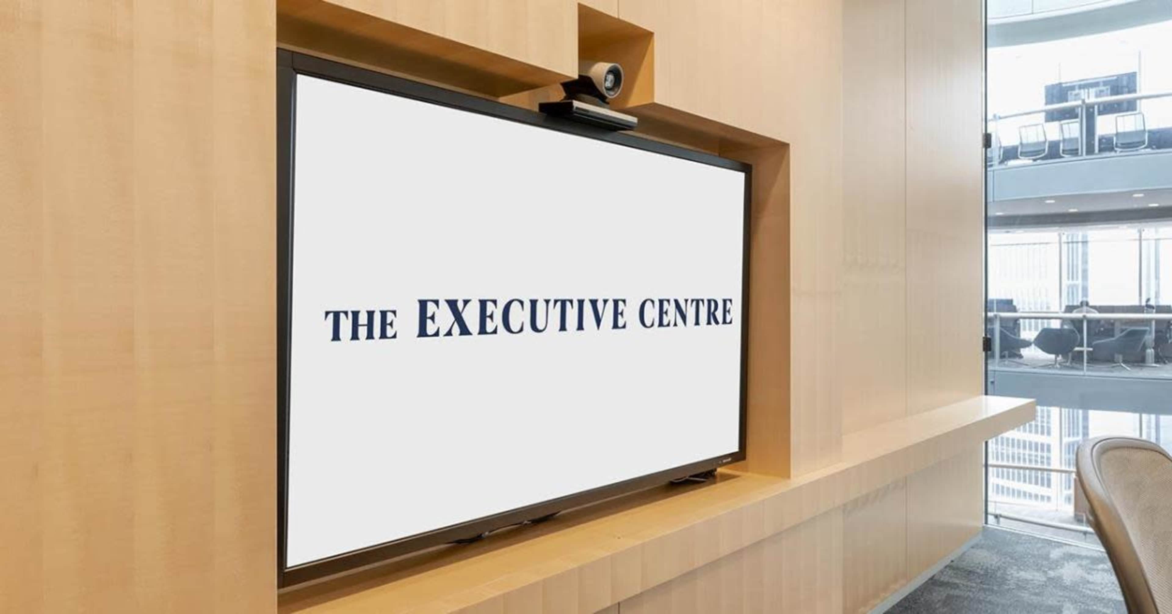 A large television displaying the text "THE EXECUTIVE CENTRE" is mounted on a wooden wall in a modern office space.