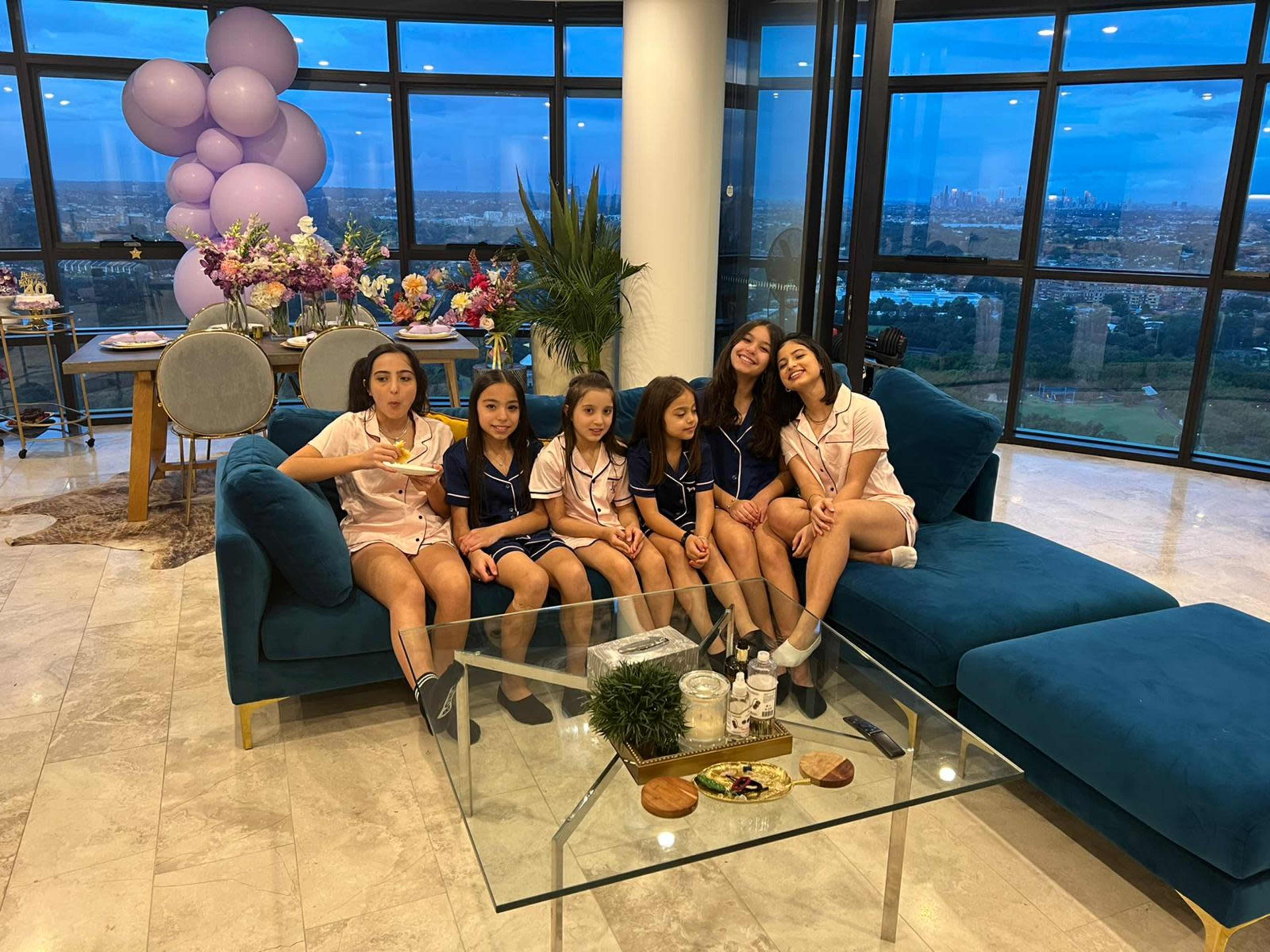 A group of six children in matching pajamas poses smiling on a sofa in a bright, modern living room with large windows and decorative elements.