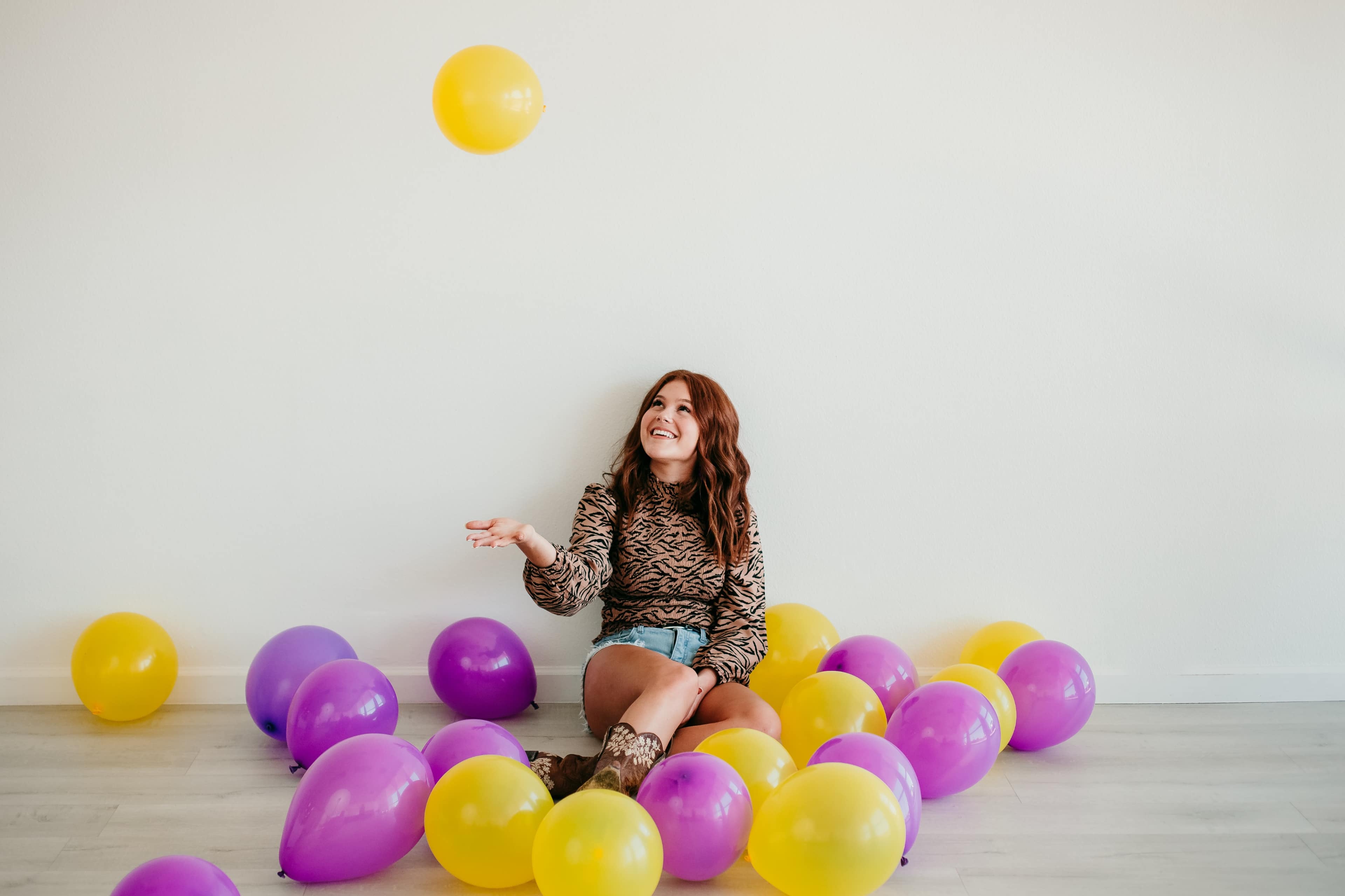 A woman sits on the floor surrounded by purple and yellow balloons, playfully tossing a yellow balloon into the air.