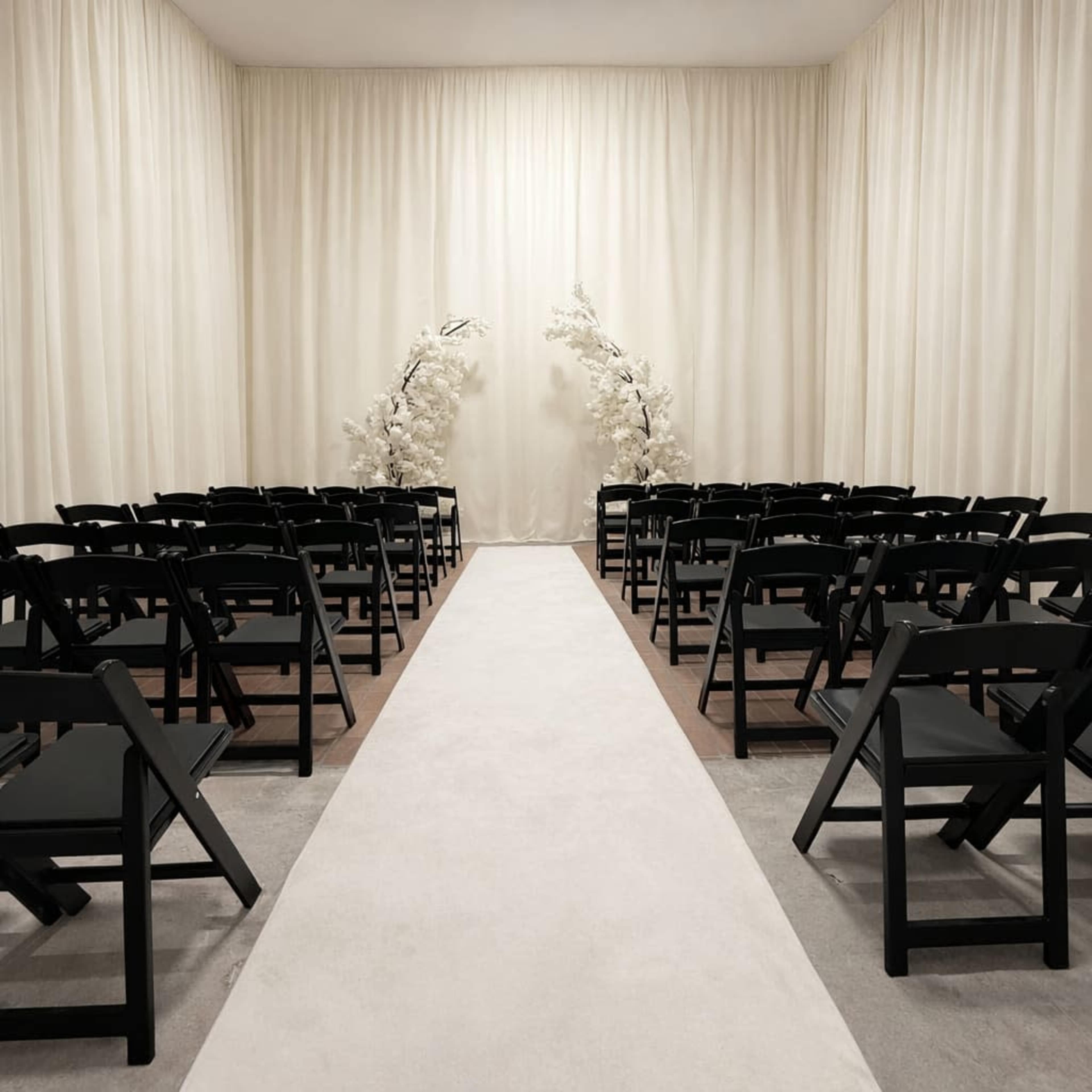 The image shows a wedding ceremony setup with rows of black chairs facing a central aisle, flanked by white floral arrangements against a backdrop of draped cream fabric.