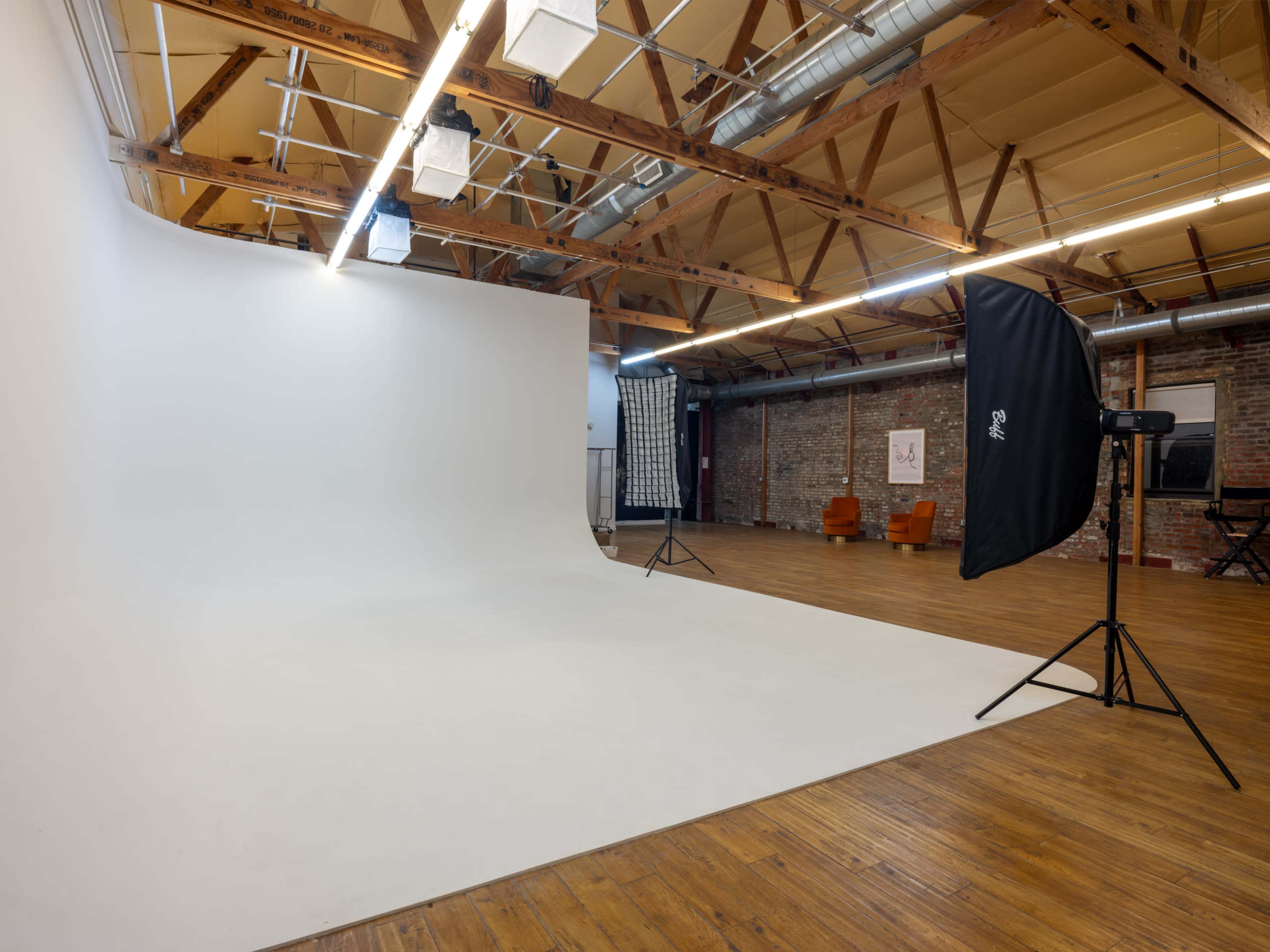 The image shows a spacious photography studio with a large white seamless backdrop, wooden floors, exposed beams, and a few brown chairs in the background.