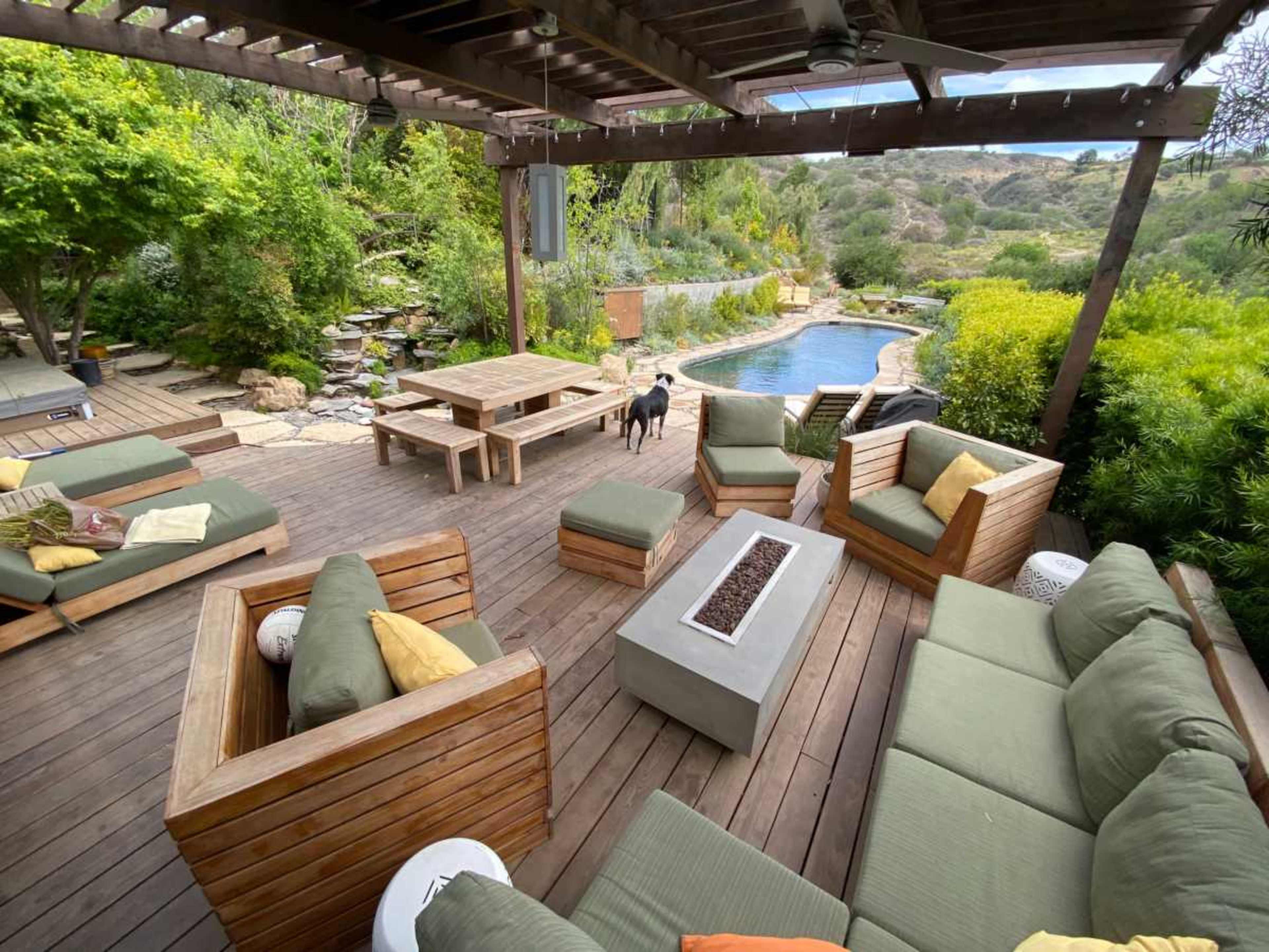The image shows a wooden deck with seating areas, a fire pit, and a swimming pool surrounded by greenery.
