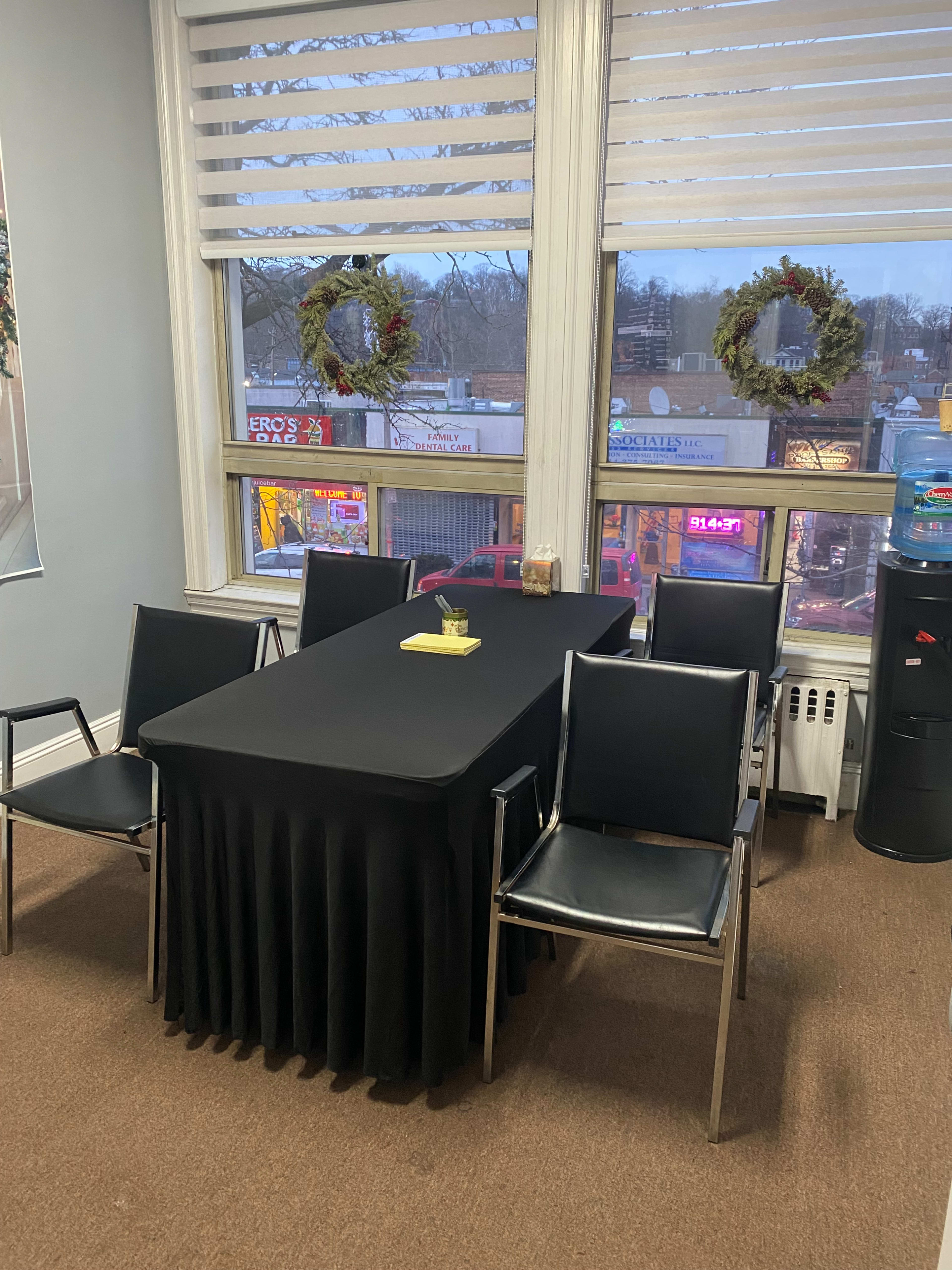 A small meeting area features a table covered with a black cloth and six black chairs facing a window with wreaths, overlooking a street lined with shops.