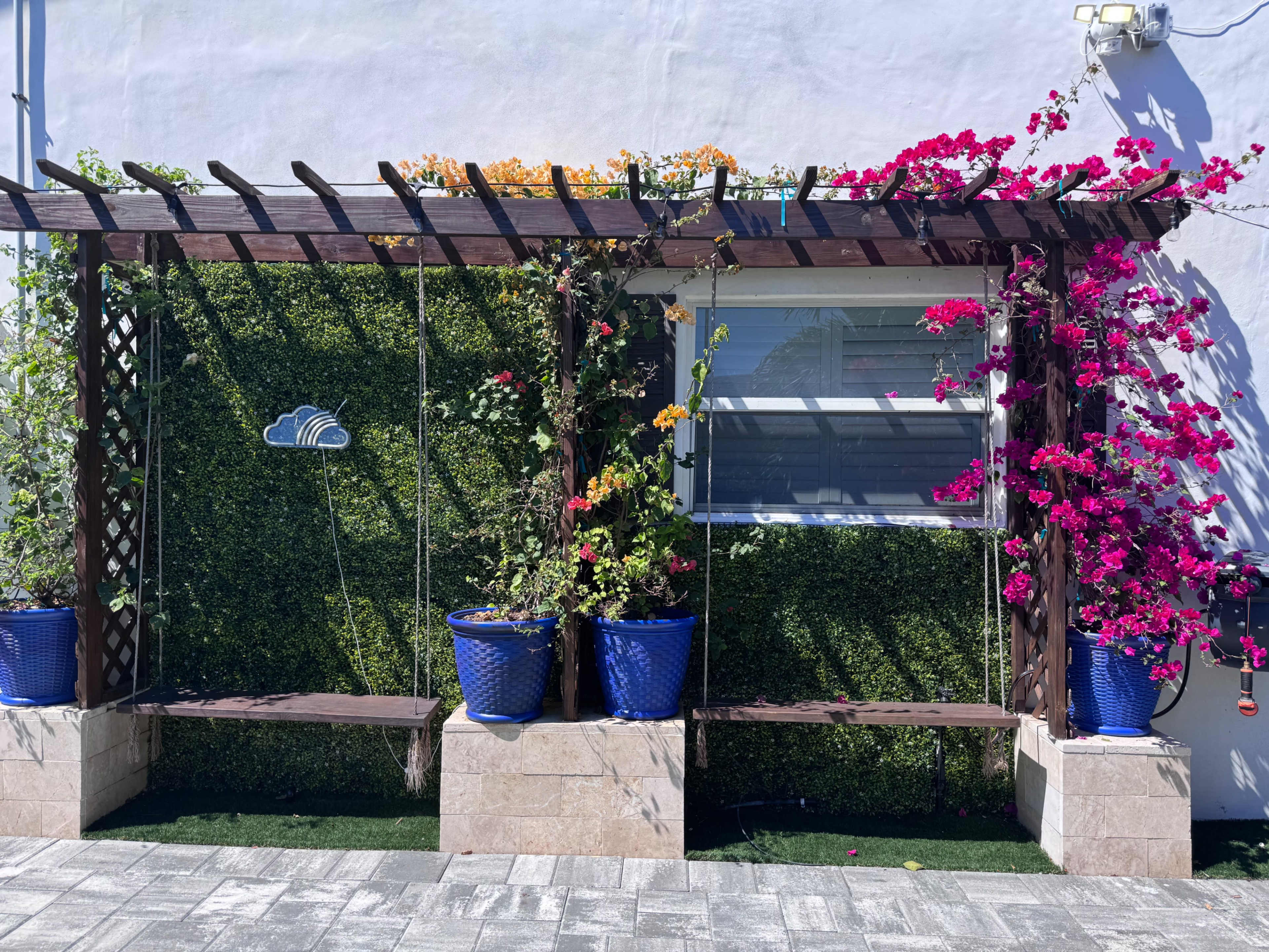 The image shows a wooden pergola adorned with colorful bougainvillea plants, flanking a window and featuring two blue pots on a tiled patio.