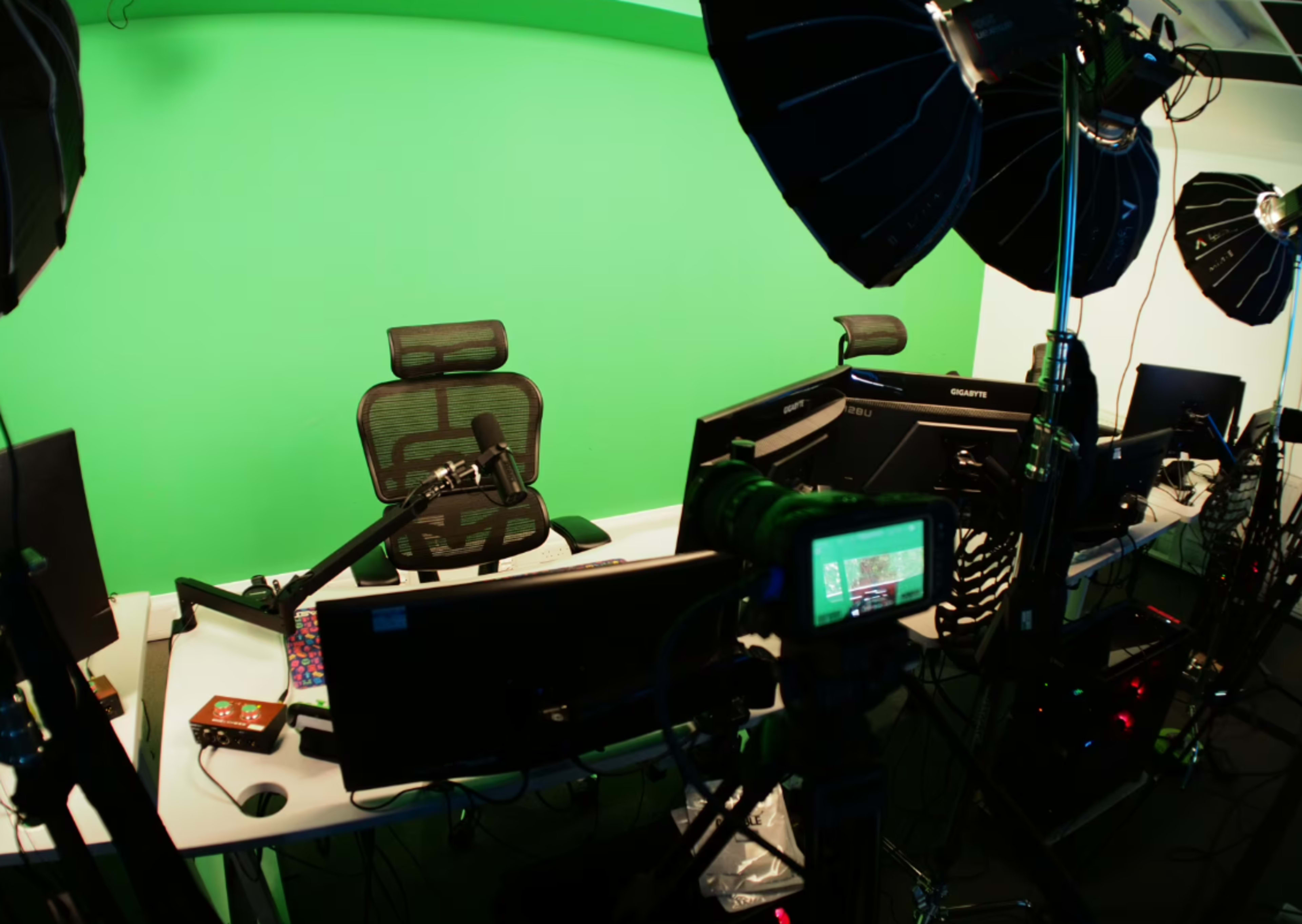 The image shows a recording setup with multiple computers, a microphone, and studio lighting in front of a green screen.