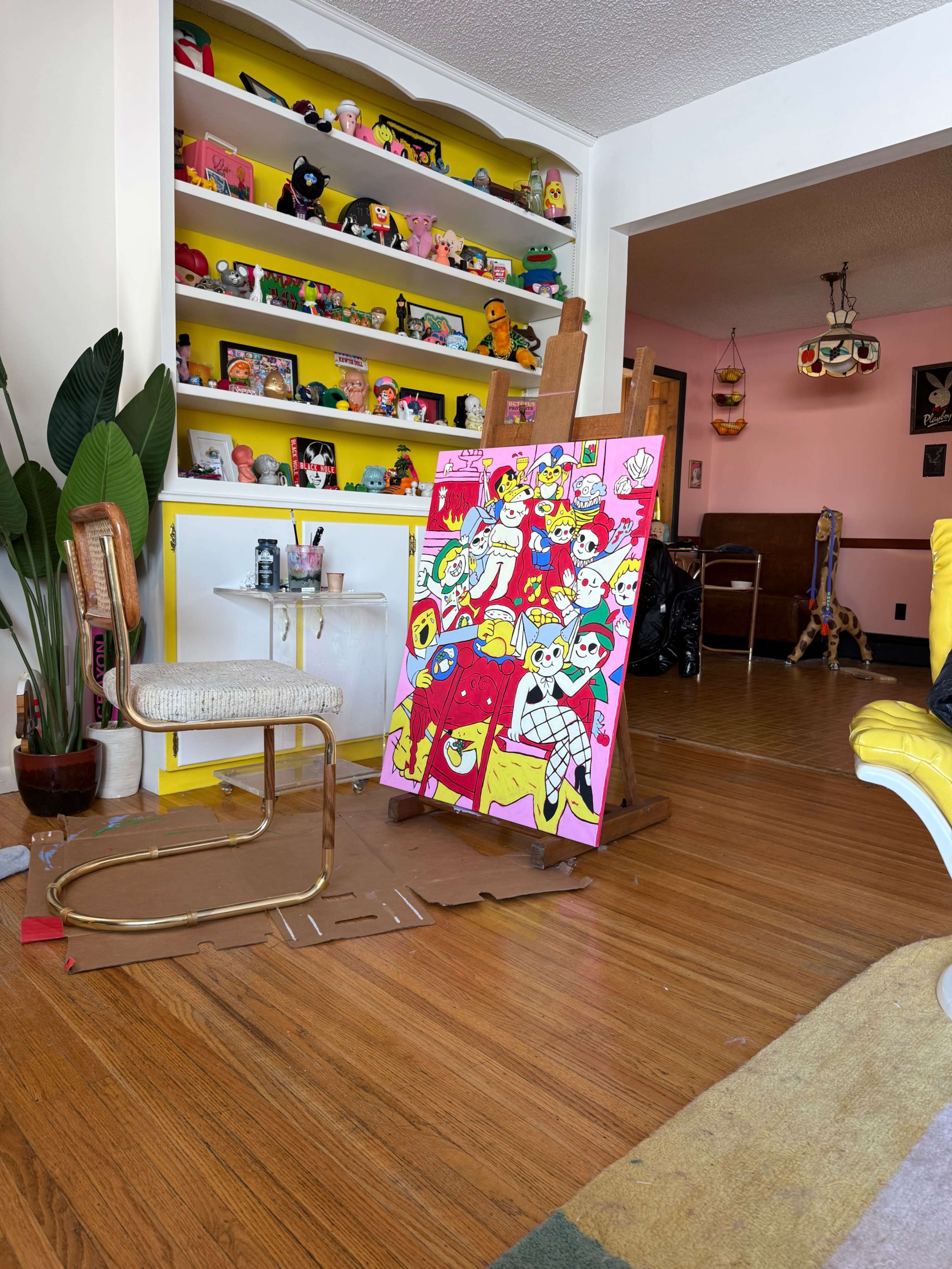 The image shows a colorful room with a bright pink wall displaying an art piece on an easel, surrounded by various decorative items and plants.