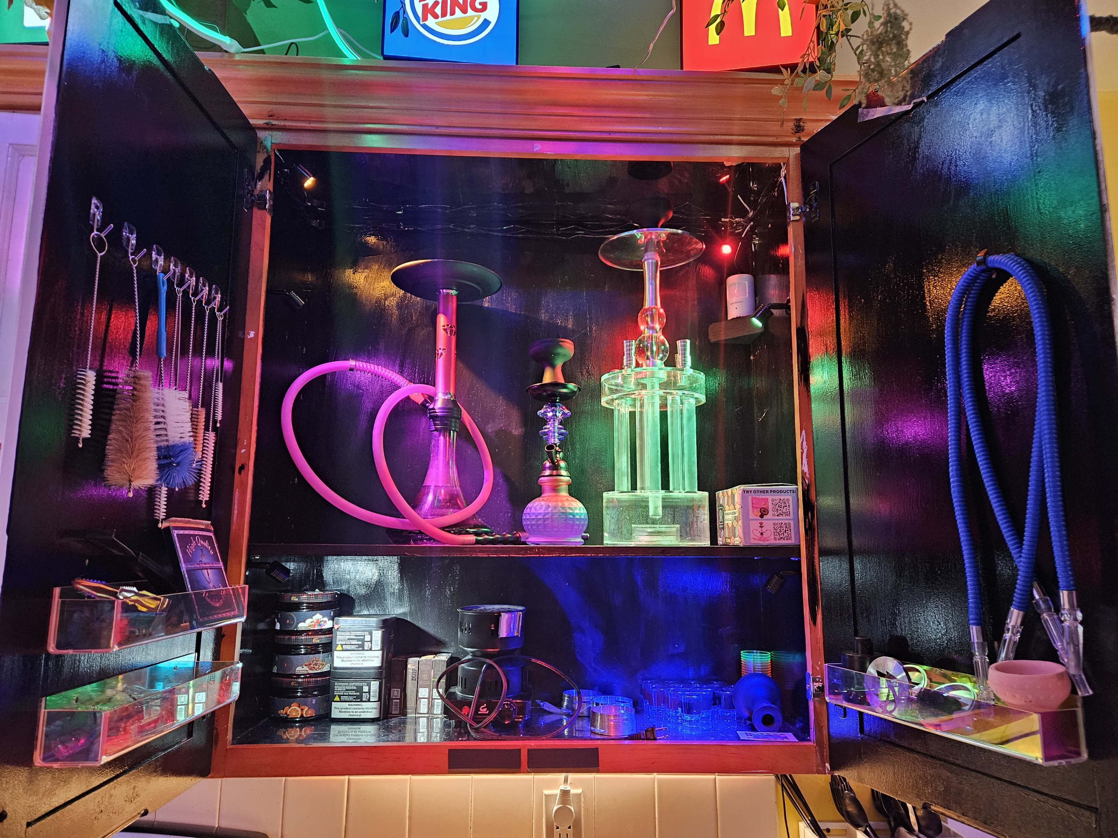 The image shows a wooden cabinet filled with various hookah accessories, including hoses, bowls, and cleaning tools, illuminated by colorful lights.