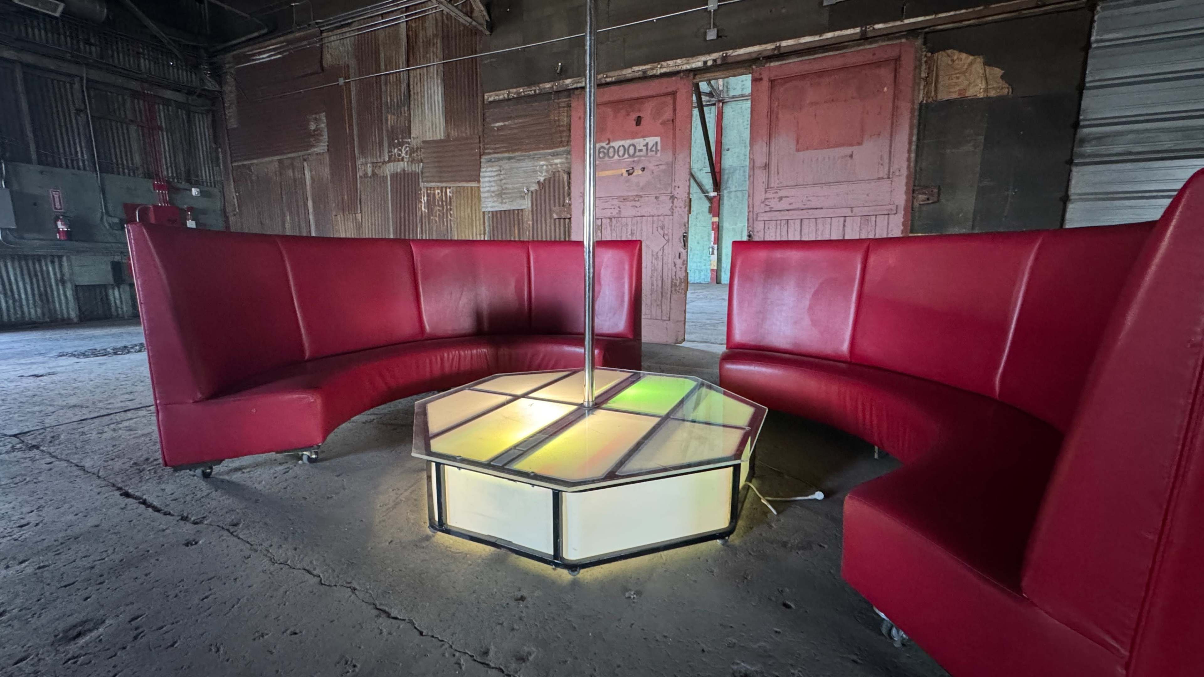 The image shows a circular seating arrangement made of red vinyl-backed booths surrounding a hexagonal lighted table in an industrial space with weathered walls and doors.