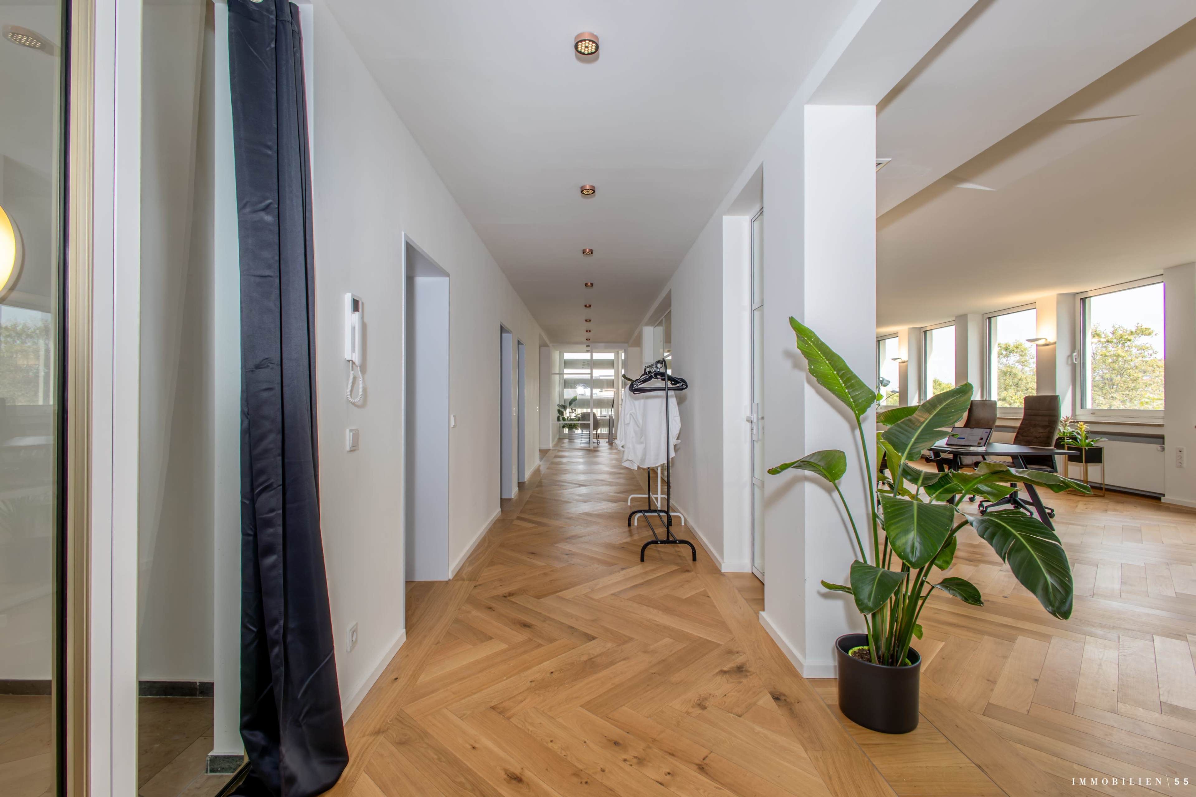 The image shows a long, well-lit hallway with wooden flooring, leading to a set of large windows, and includes coat racks and potted plants along the sides.