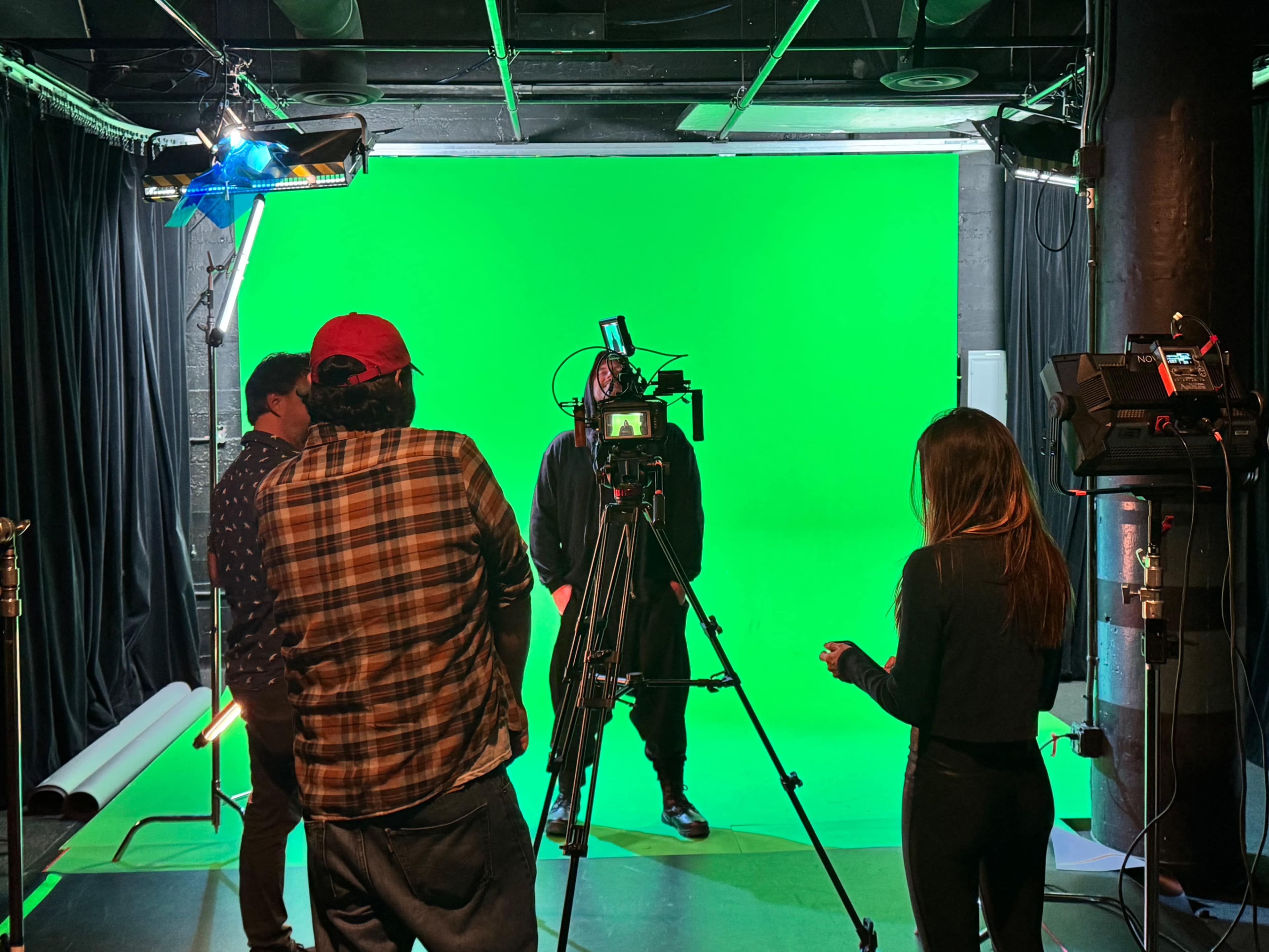 A film crew sets up equipment in front of a green screen in a studio for a shoot.