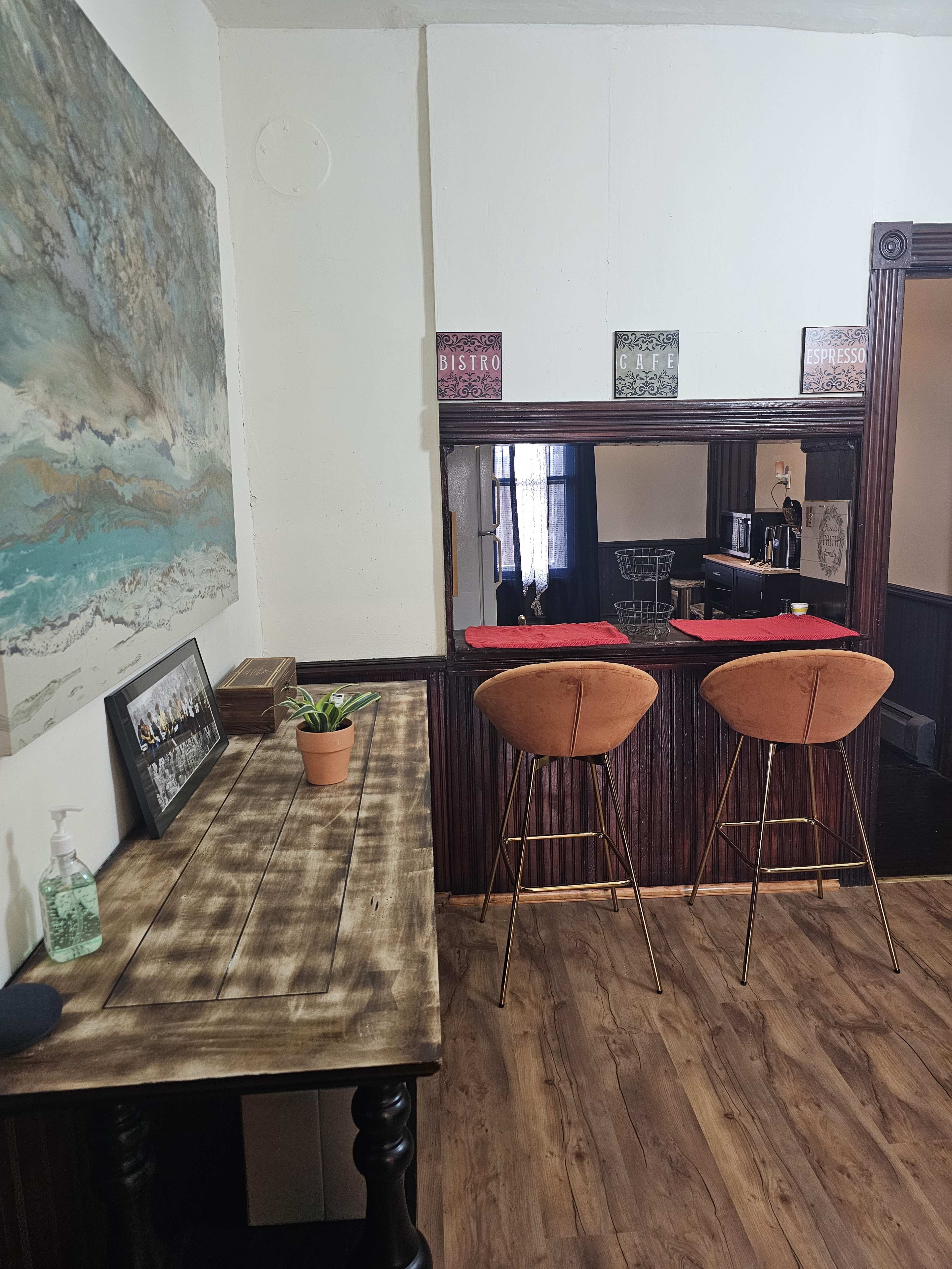 The image shows a small interior space featuring a wooden table, two bar stools at a counter, and a painting on the wall.