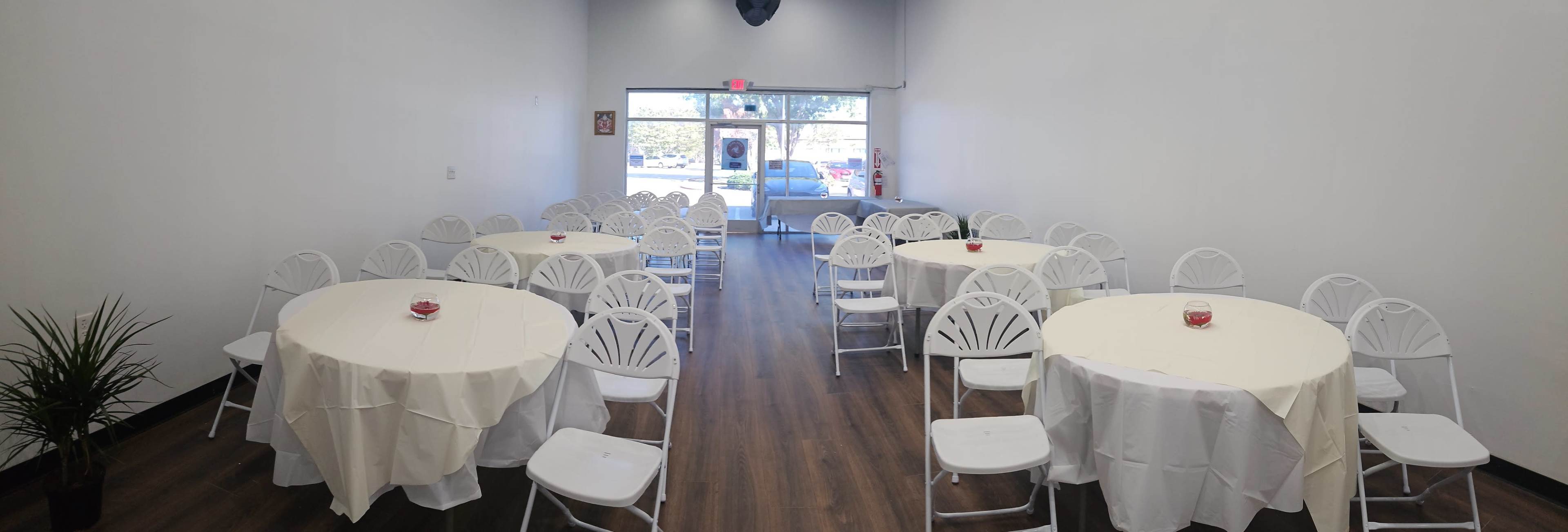 A spacious room arranged with several round tables covered in white tablecloths and surrounded by white chairs, leading to a large front window.