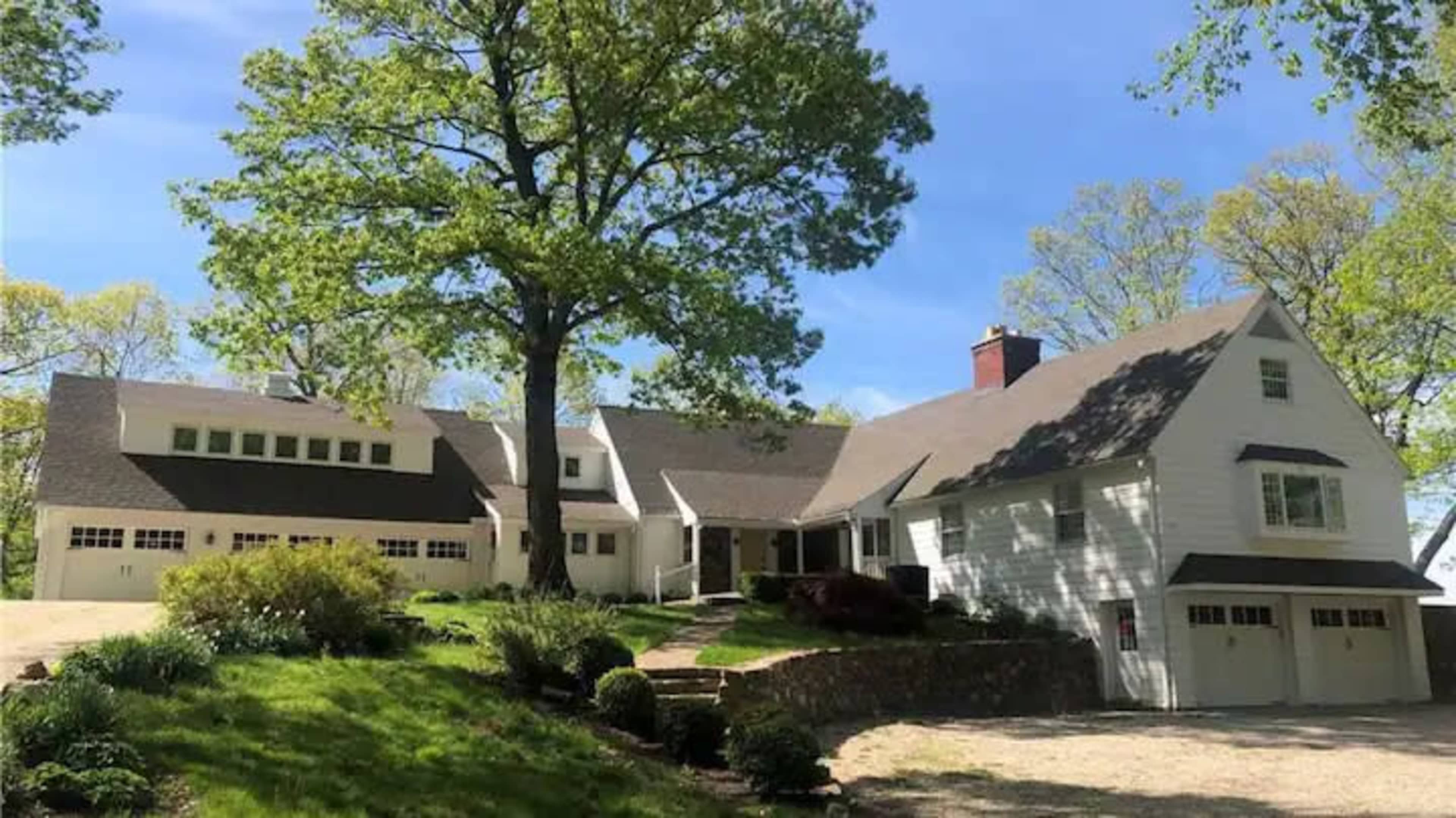 The image shows a large white house with multiple sections, situated on a landscaped property with a driveway and large trees.