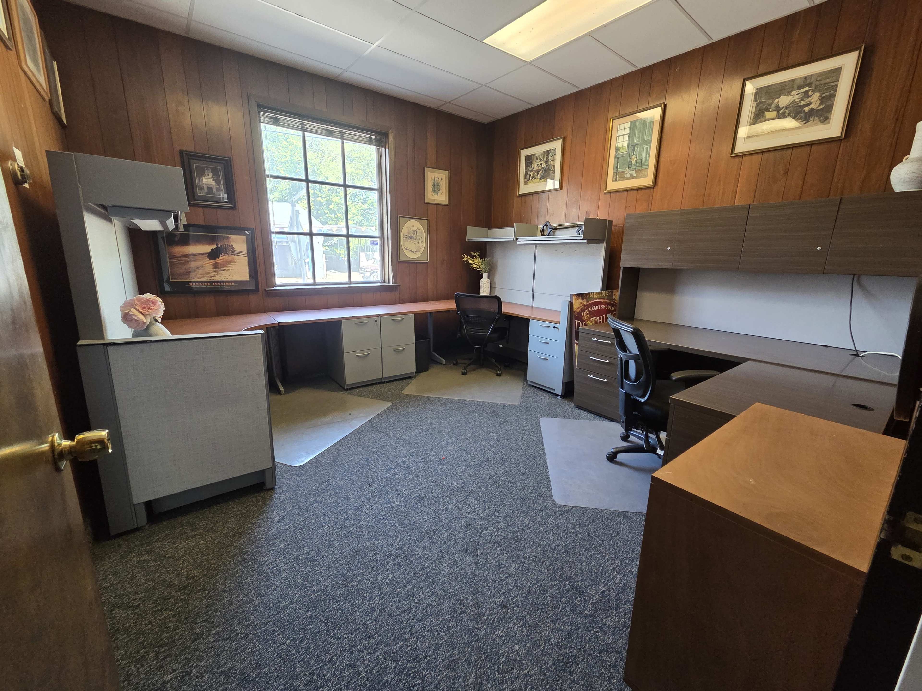 The image shows a tidy office space with wooden paneling, a large window, two desks, an office chair, and framed artwork on the walls.