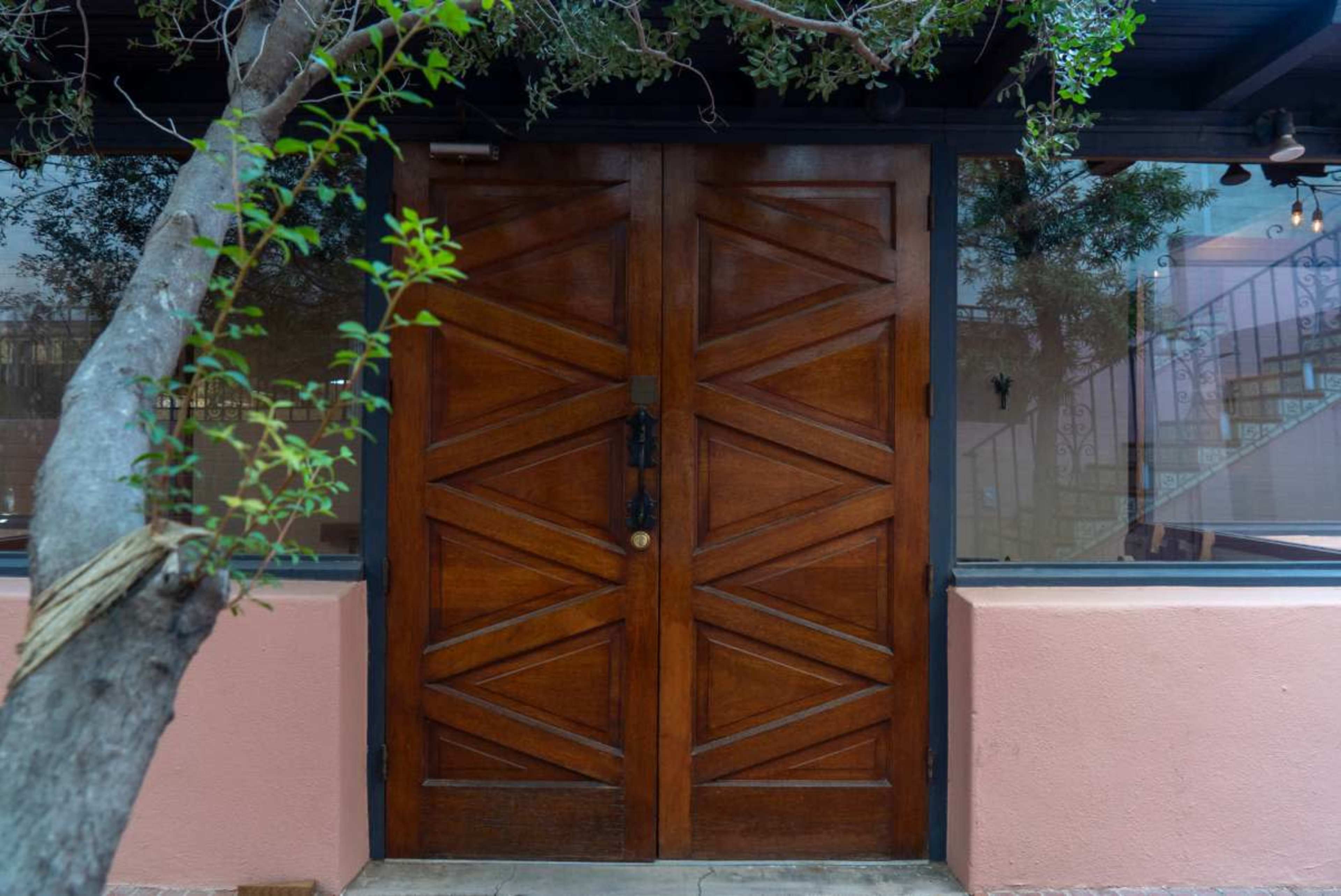 The image shows a large wooden door with a distinctive chevron pattern, framed by a pink wall and surrounded by greenery.