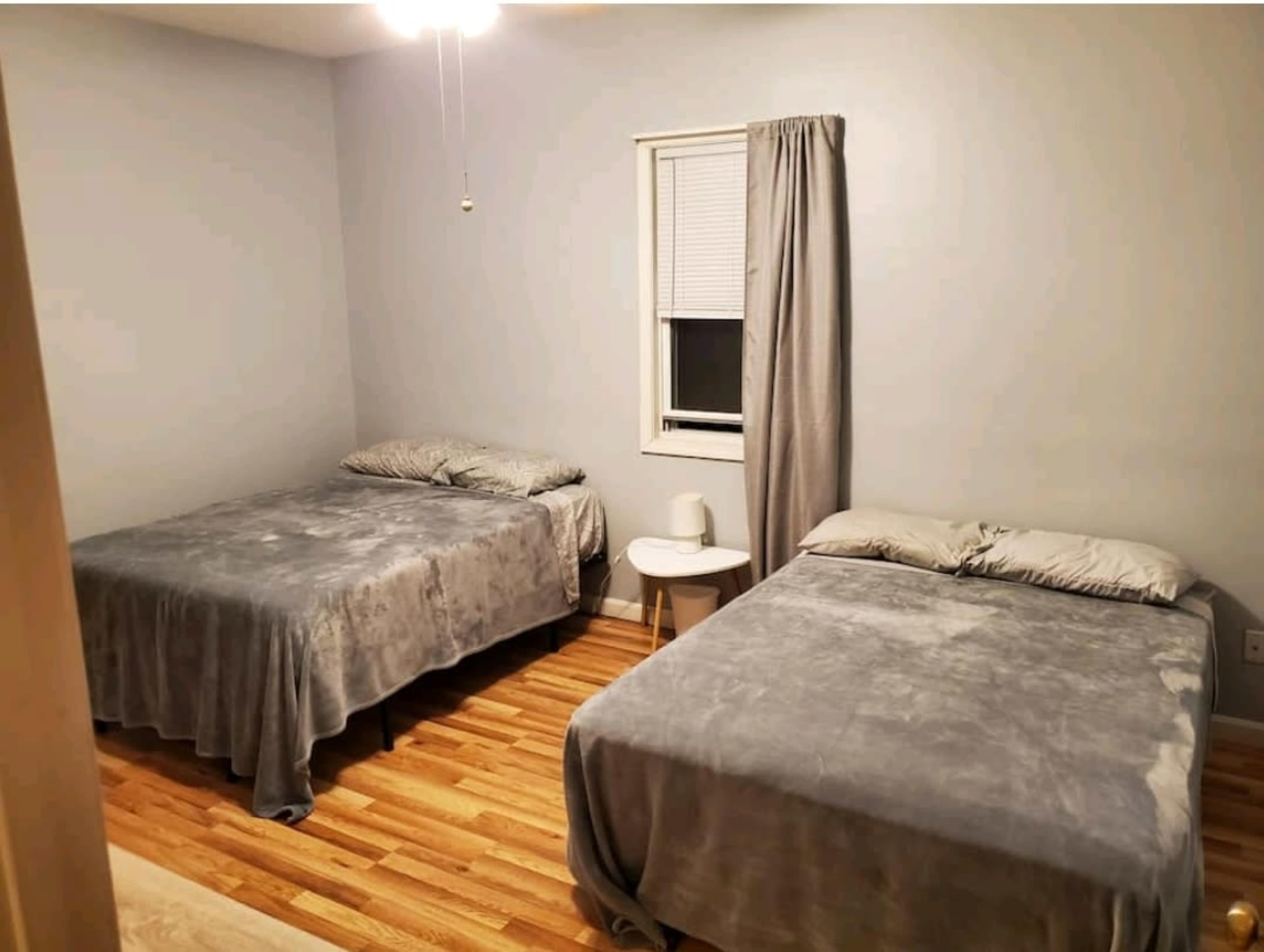 A simple bedroom with two beds covered in gray blankets, a small bedside table, and a window with a light curtain.