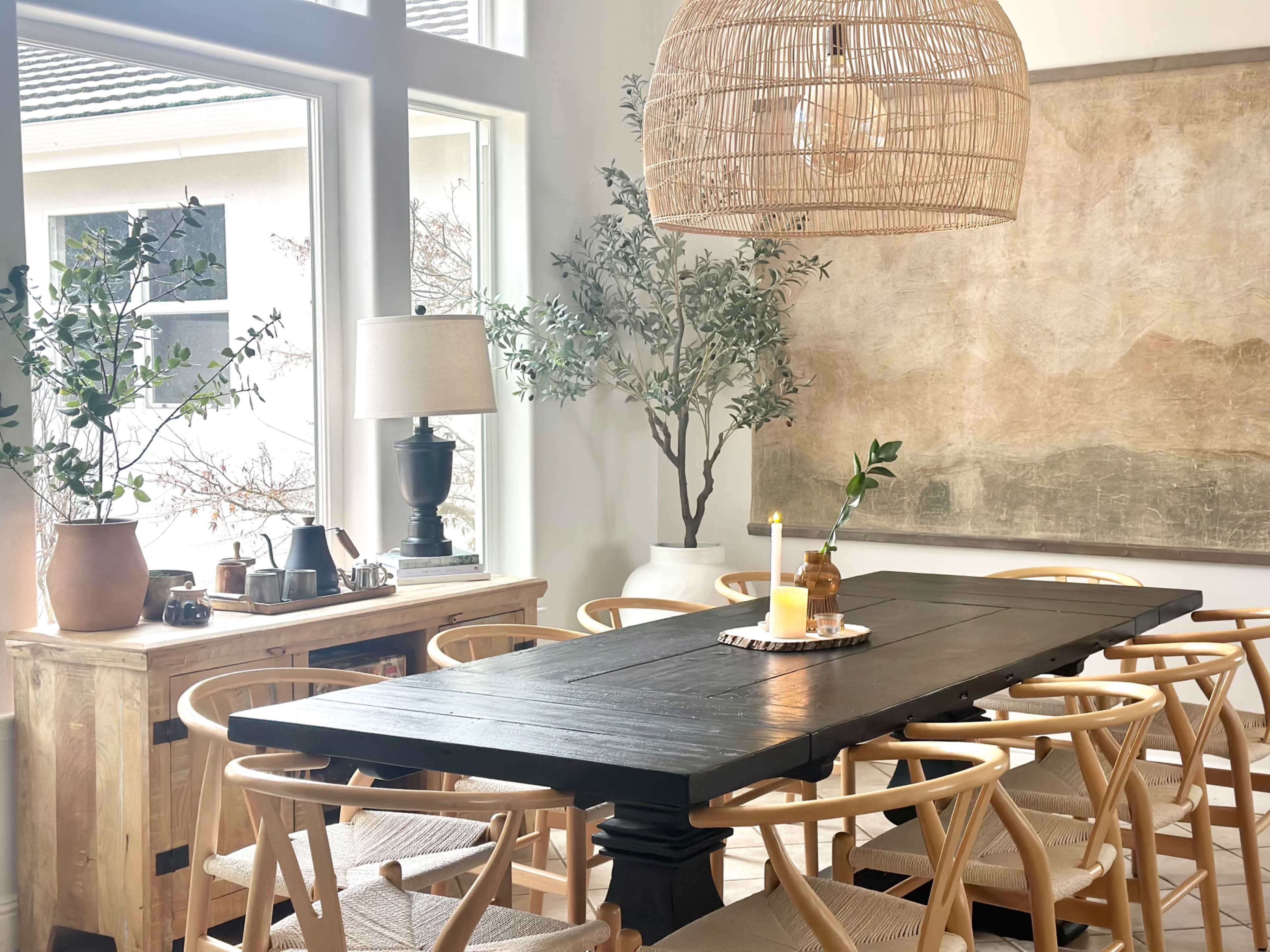 A large wooden dining table is set in a well-lit room with natural decor, including potted plants and a woven chandelier above.