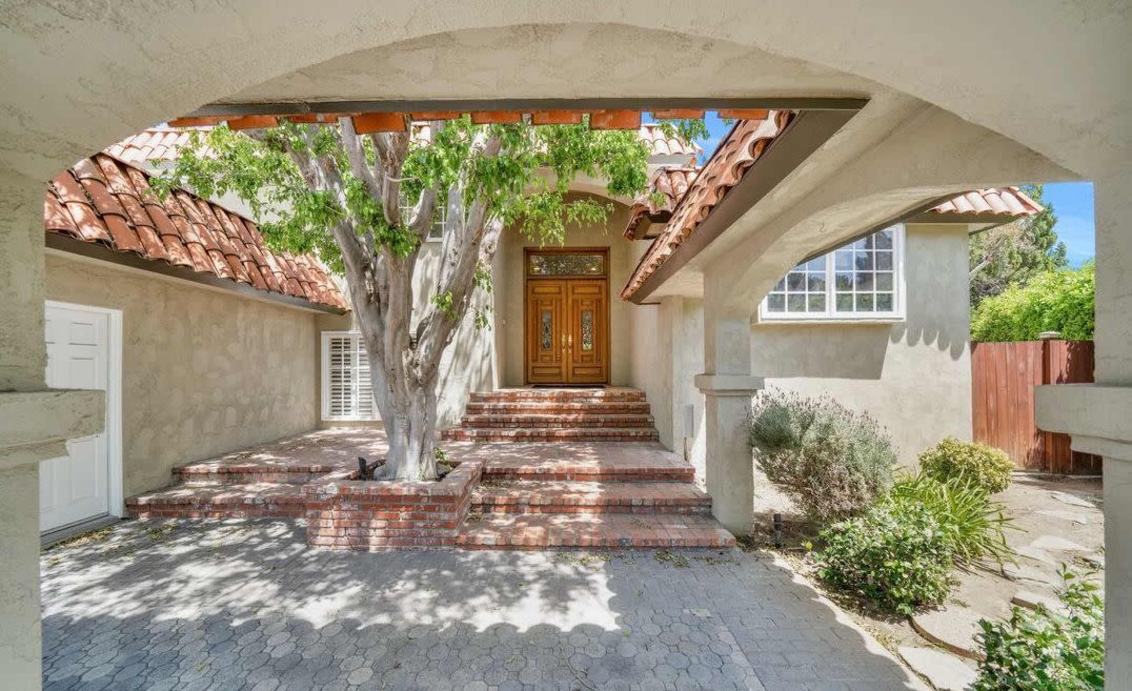 The image shows a house with a terracotta-tiled roof, a large wooden front door, and a pathway leading to the entrance framed by a tree and landscaped areas.