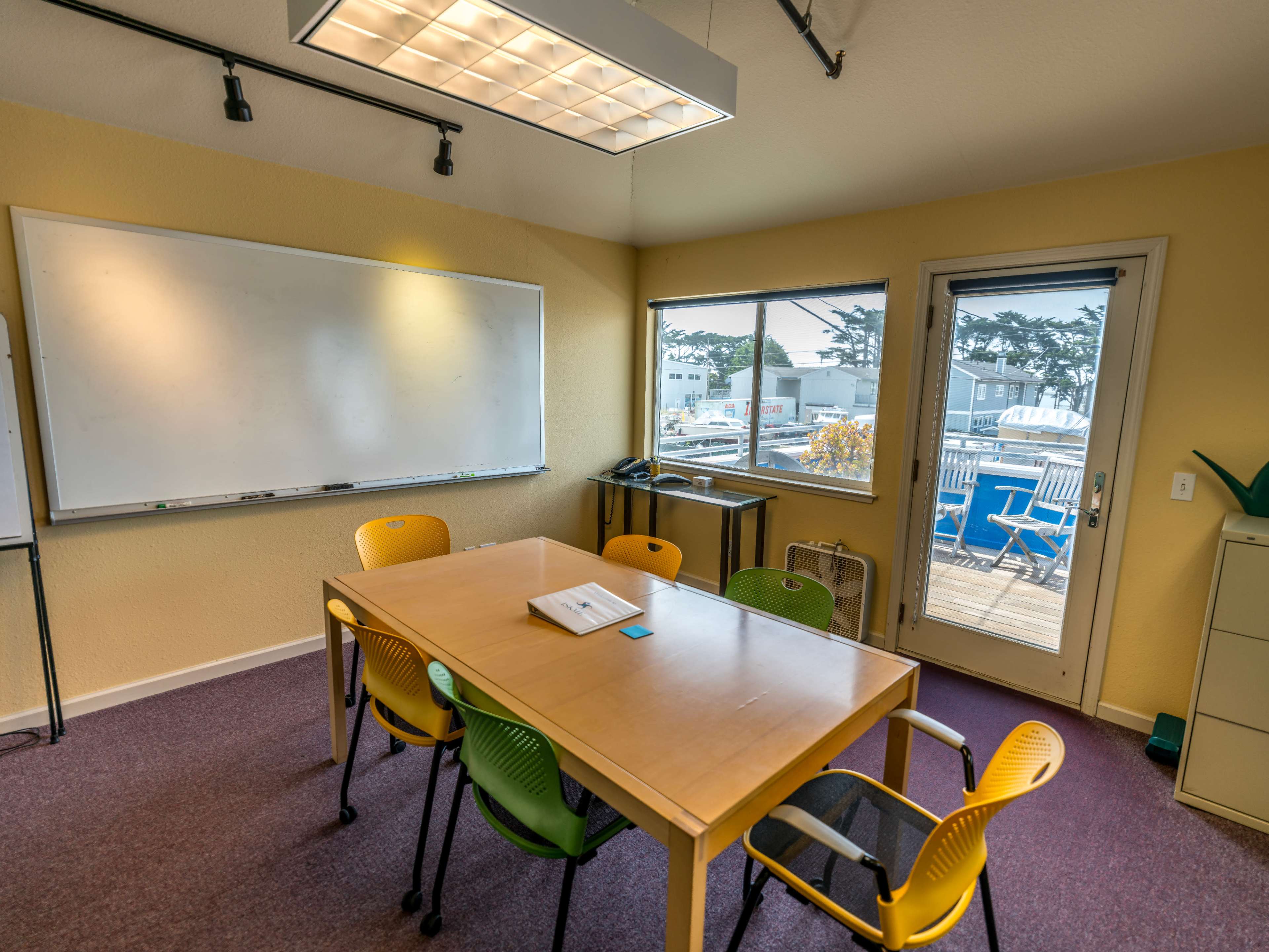 A conference room features a wooden table surrounded by colorful chairs, a whiteboard on one wall, and a view of an outdoor area through a nearby window.