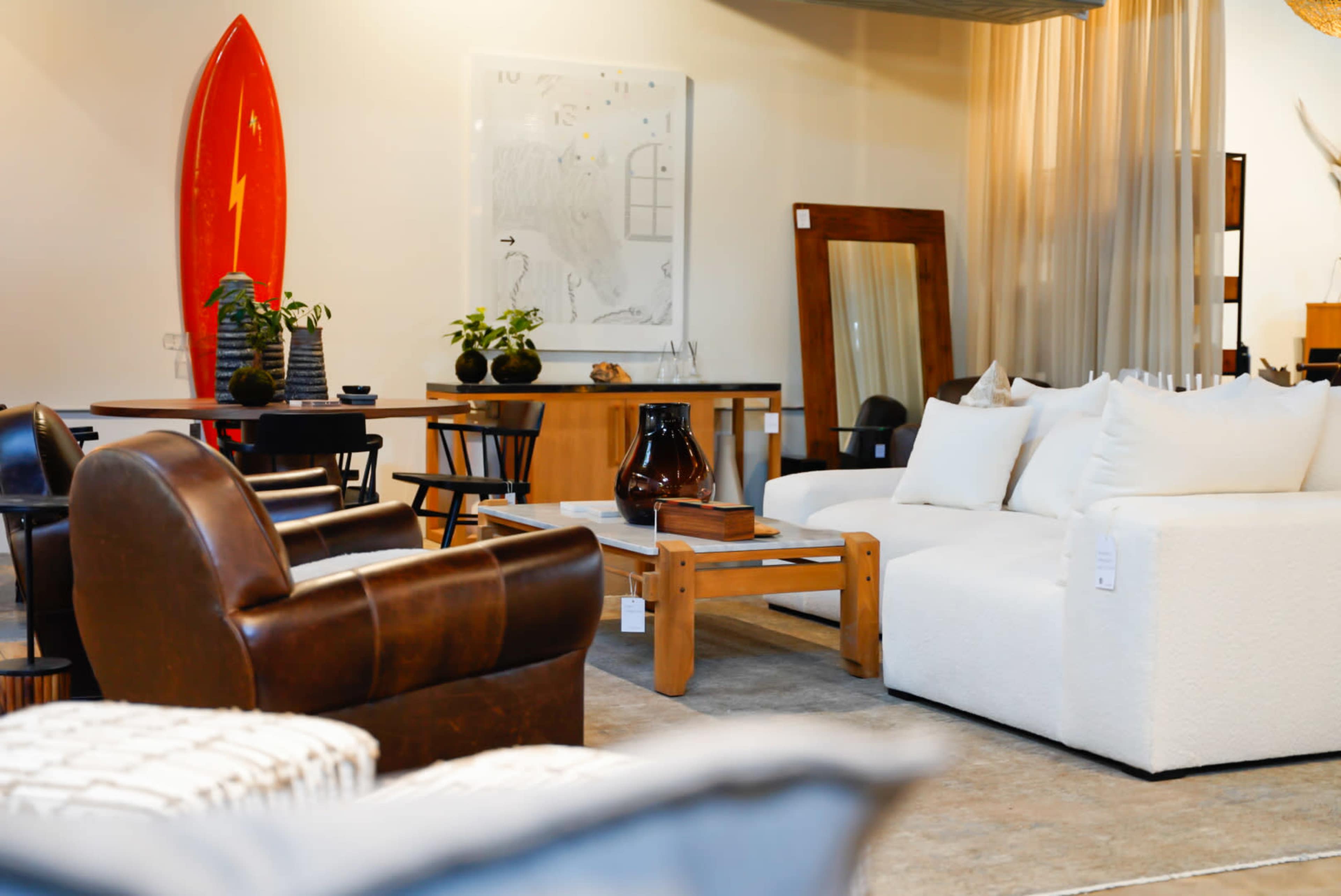 The image shows a contemporary furniture showroom featuring a variety of seating options, a wooden table, decorative plants, and a large red surfboard on display.