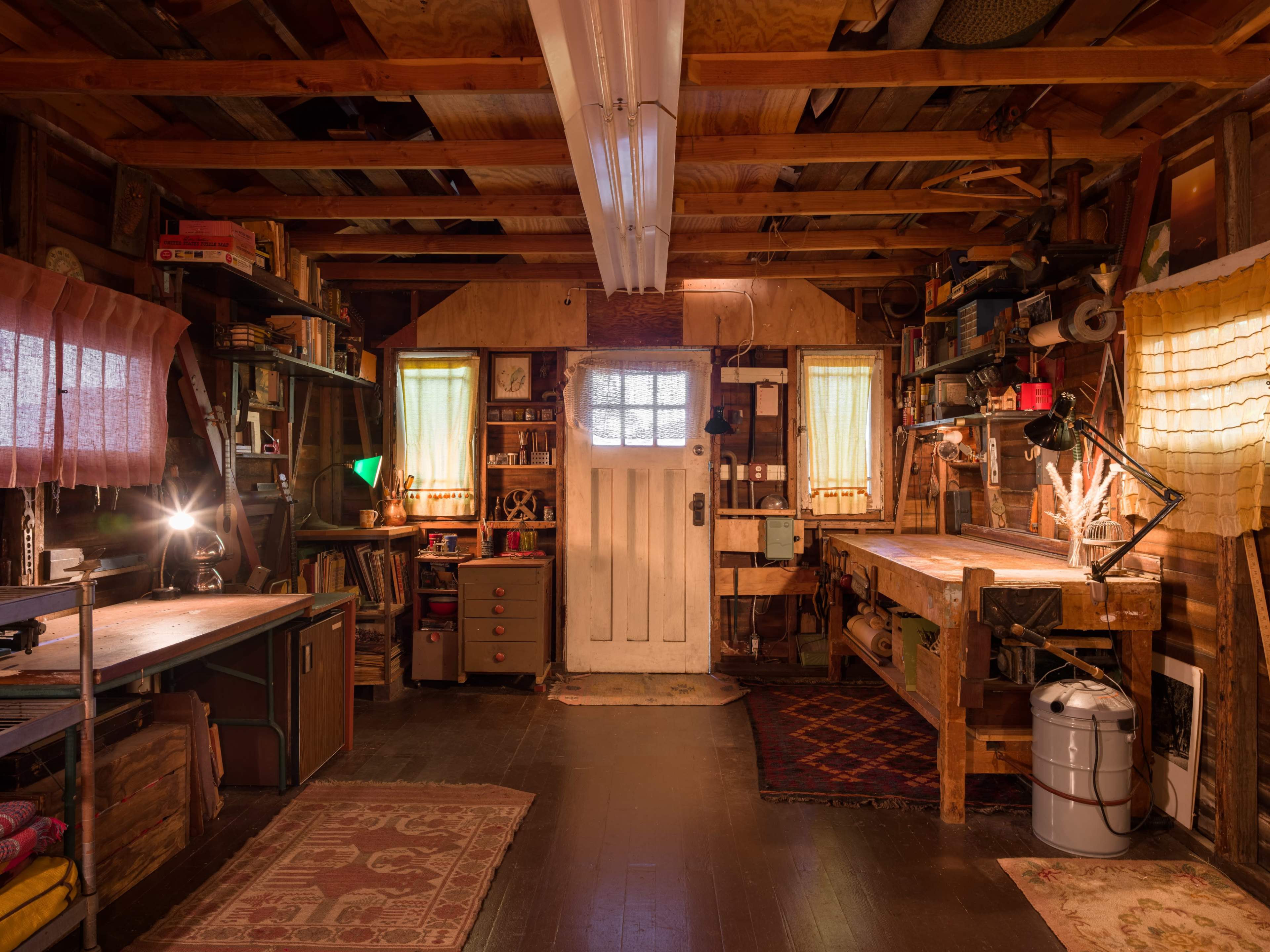 The image shows an organized workshop with wooden workbenches, shelves filled with tools and books, and a white door at the back.