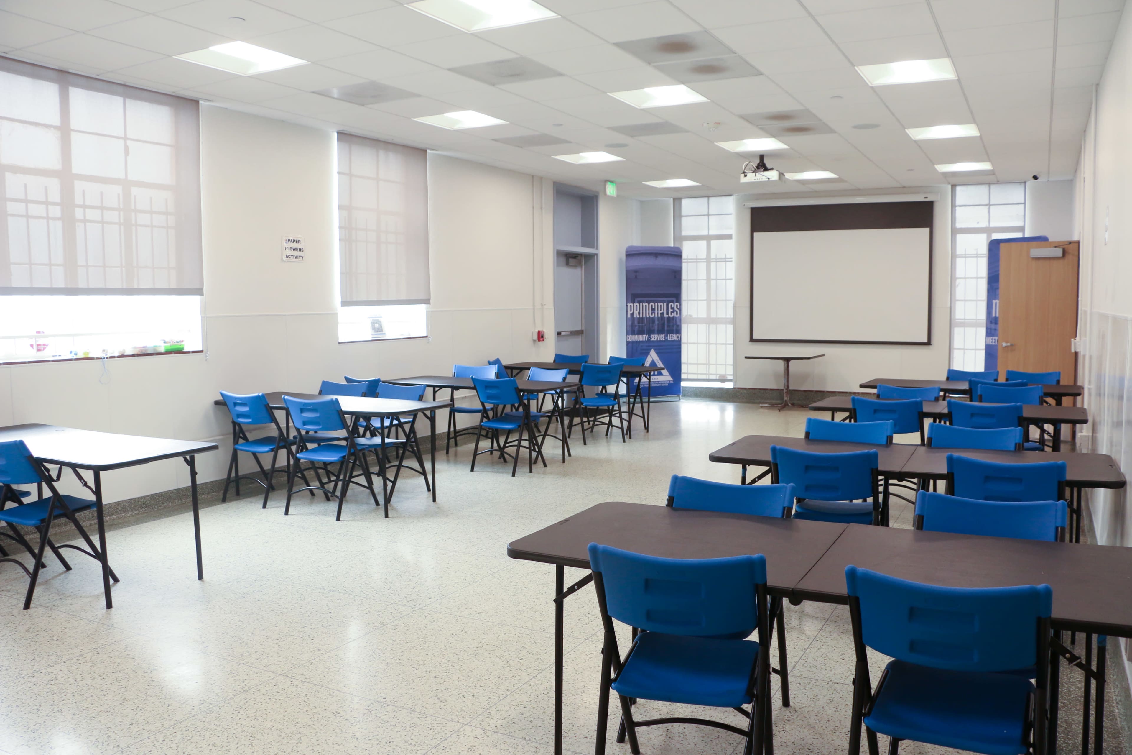 The image shows a spacious classroom with several rows of folding tables and blue chairs, equipped with a projector screen at the front and large windows letting in natural light.